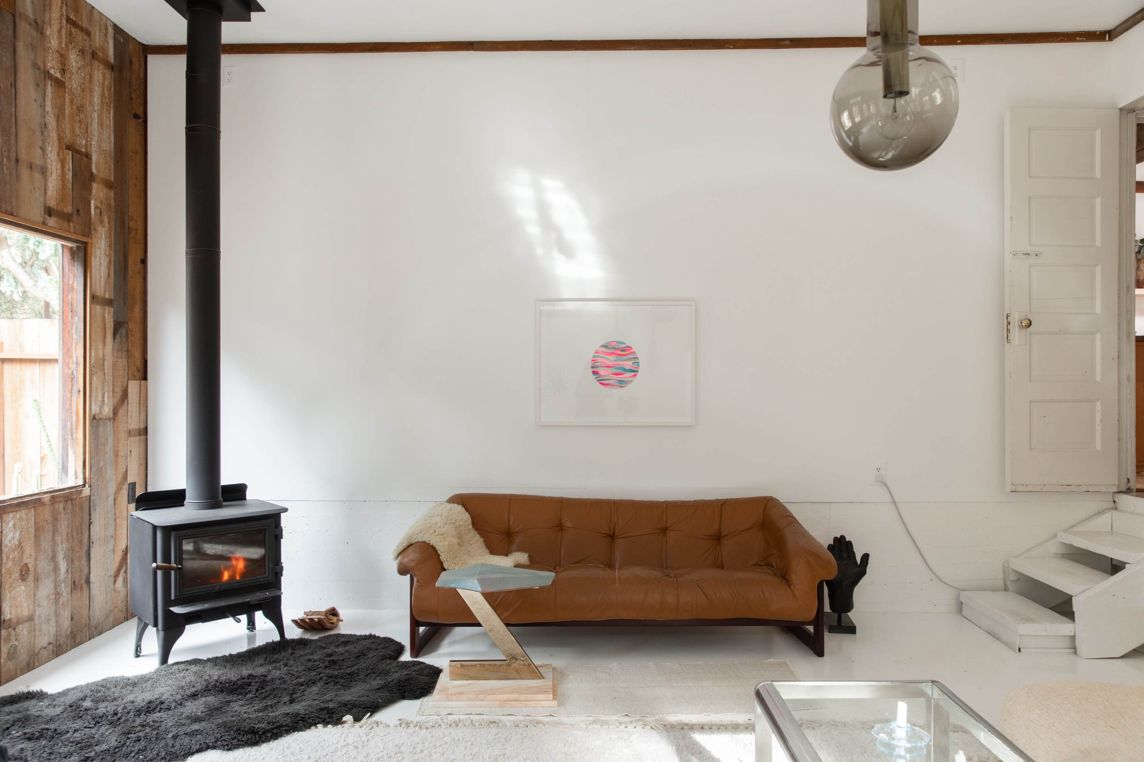 A brown leather sofa sits in a minimalist living room with a wood-burning stove, a glass side table, and a piece of artwork on the wall.