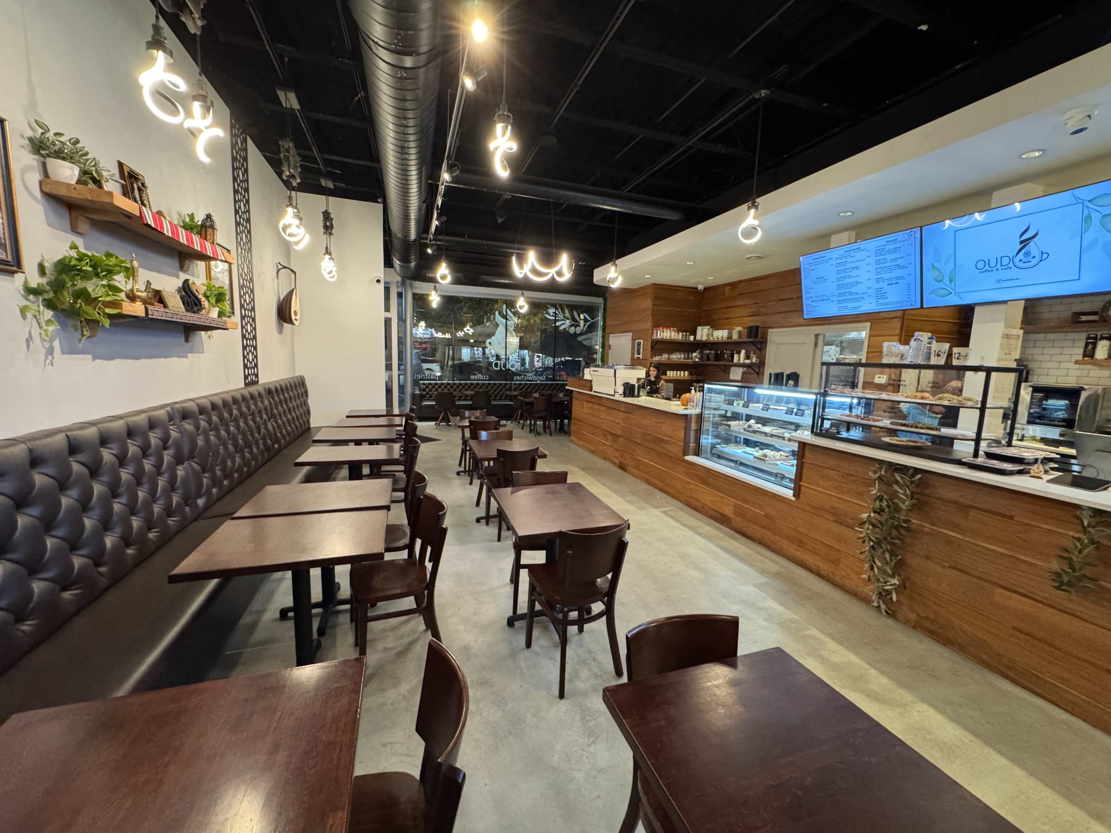 The image shows a modern café interior with wooden accents, several tables, and a display counter featuring pastries and a digital menu board.