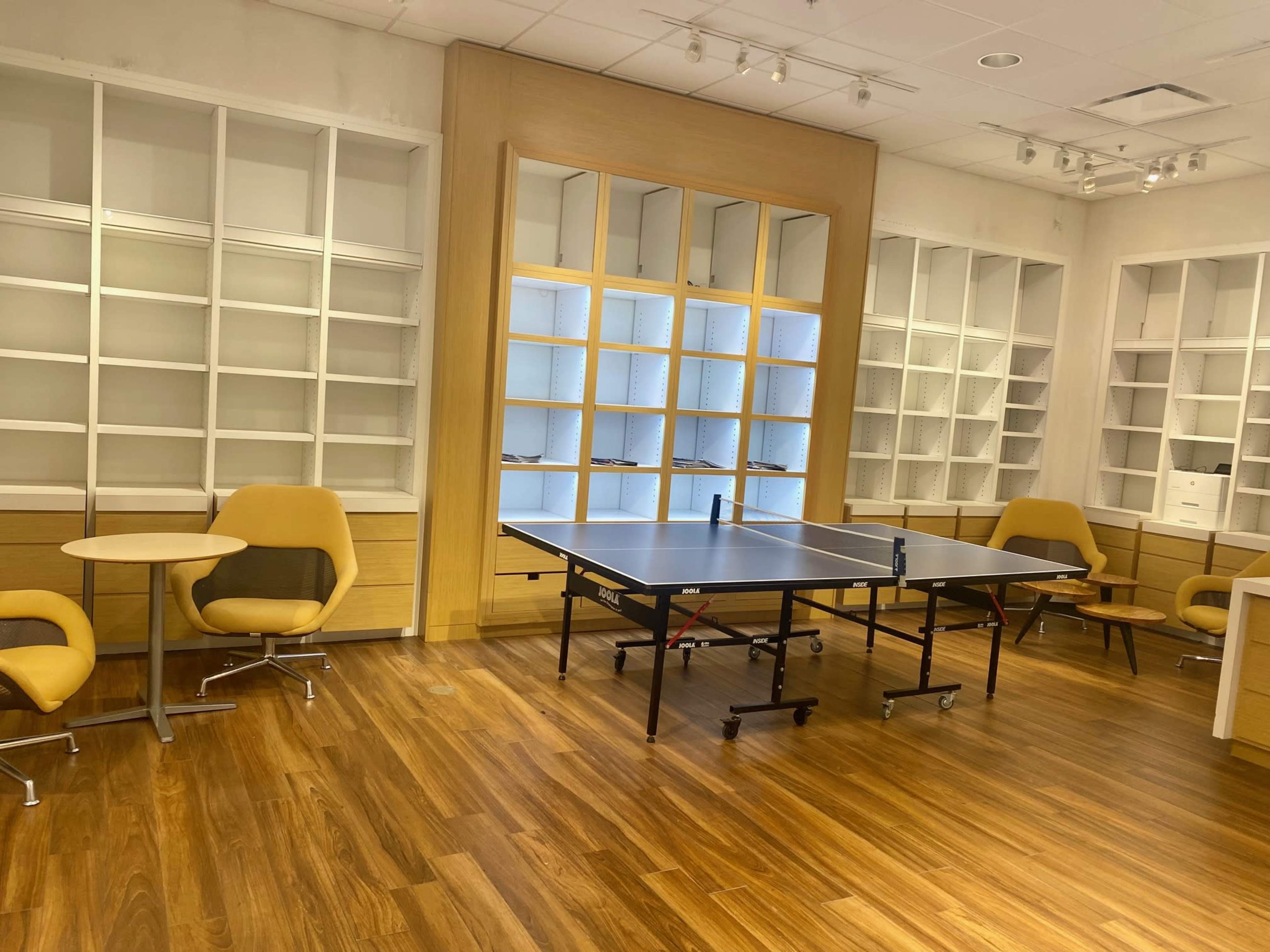 A room with wooden flooring, featuring a table tennis table in the center and two separate seating areas with chairs and tables.