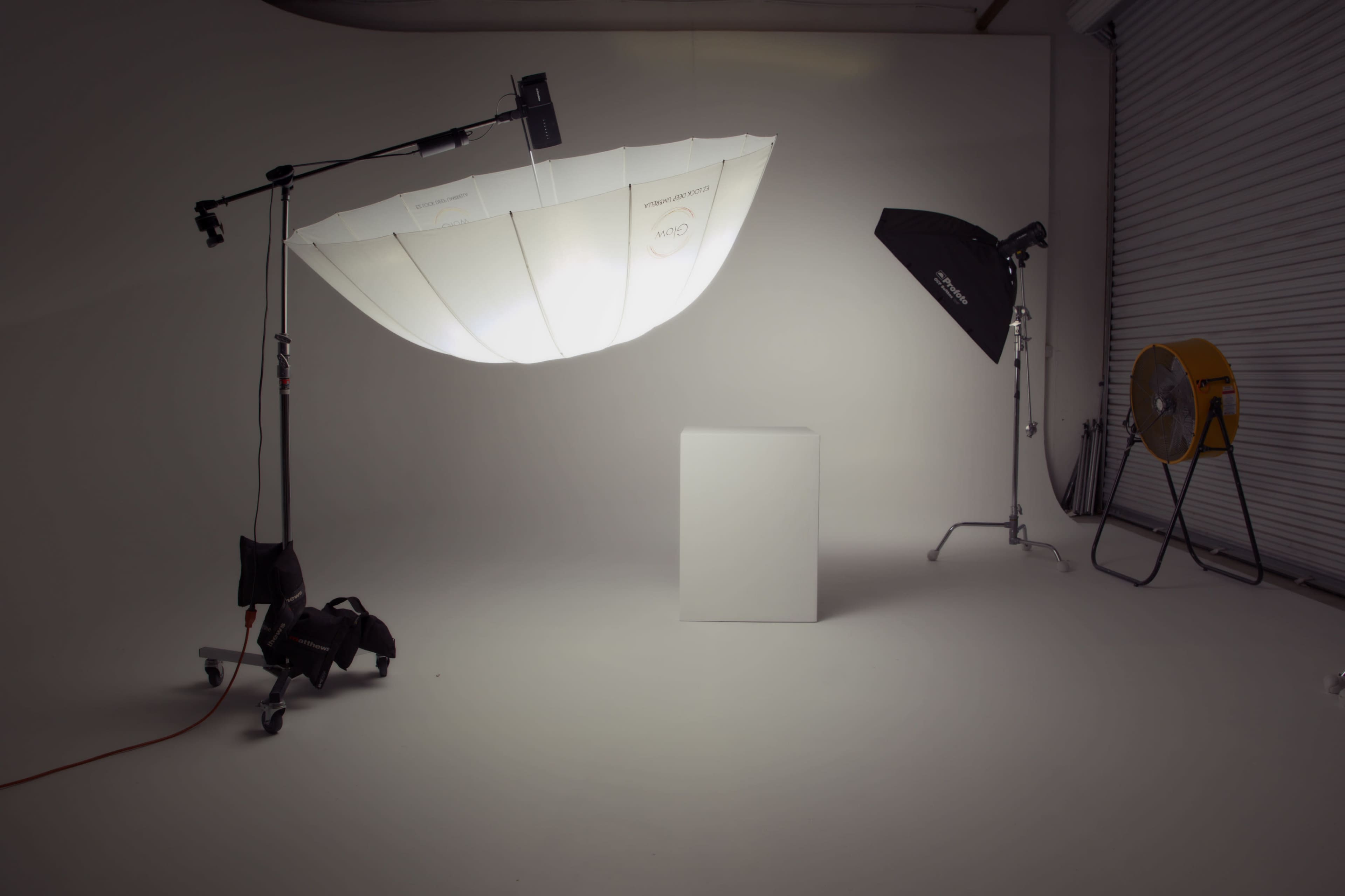 The image shows a photography studio setup with a large white umbrella light, a softbox, a fan, and a blank white pedestal on a smooth floor.