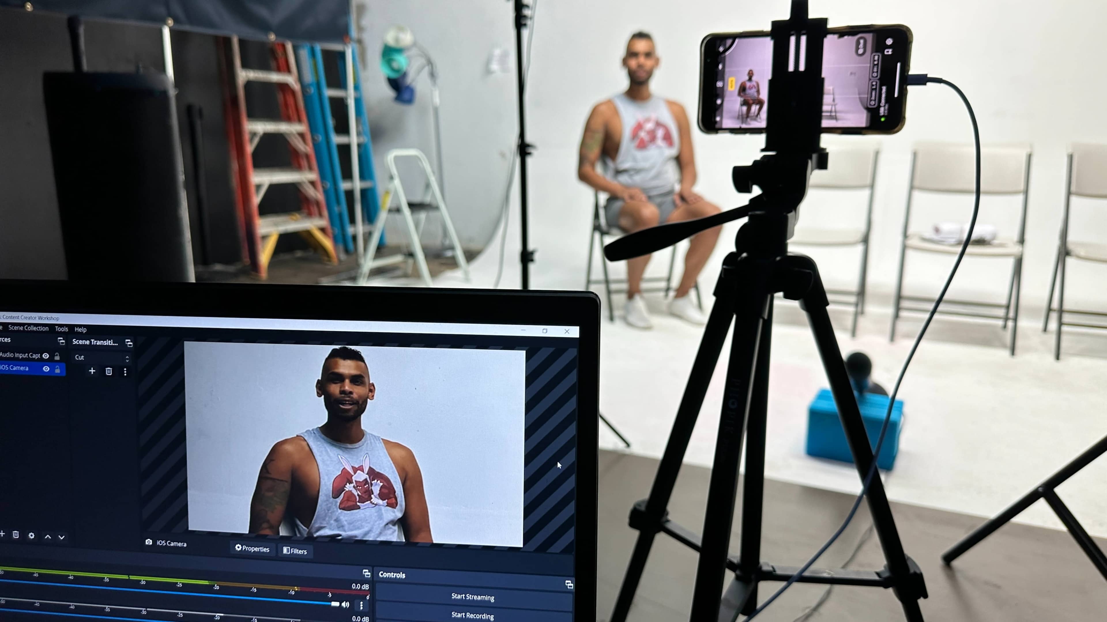 A man in a tank top sits on a white set while a camera streams the scene on a computer monitor.