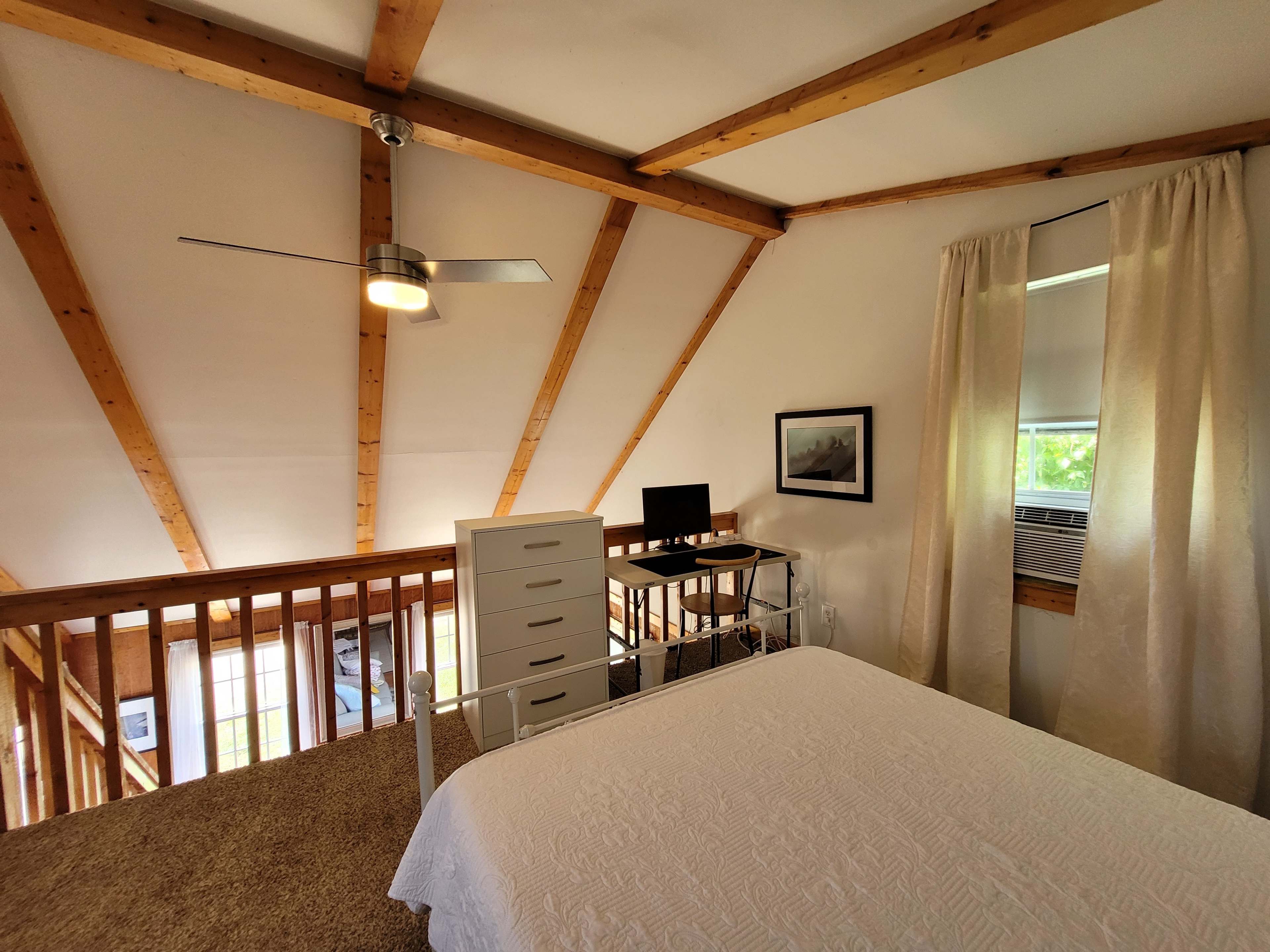 A lofted bedroom with wooden beam ceilings, a white bedspread, a dresser, and a desk by a window.
