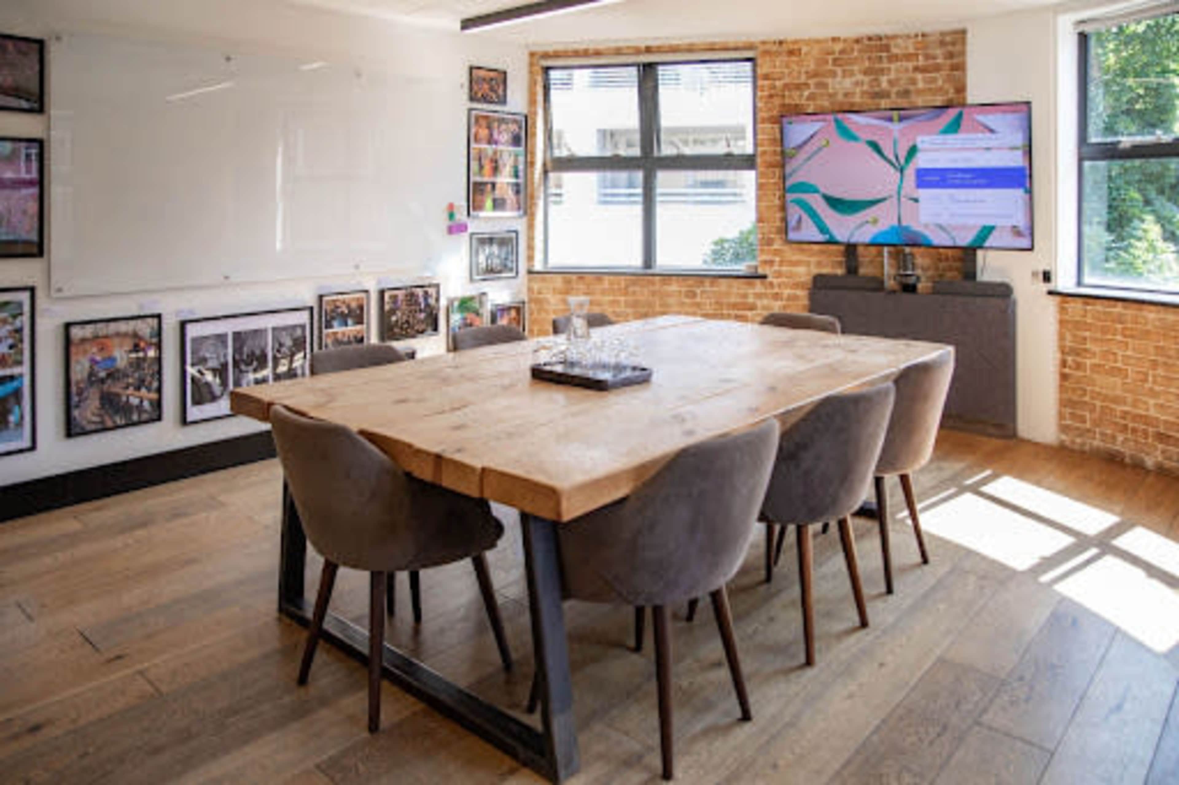 A modern conference room features a large wooden table surrounded by gray chairs, with a TV displaying content on one wall and framed photographs on another.