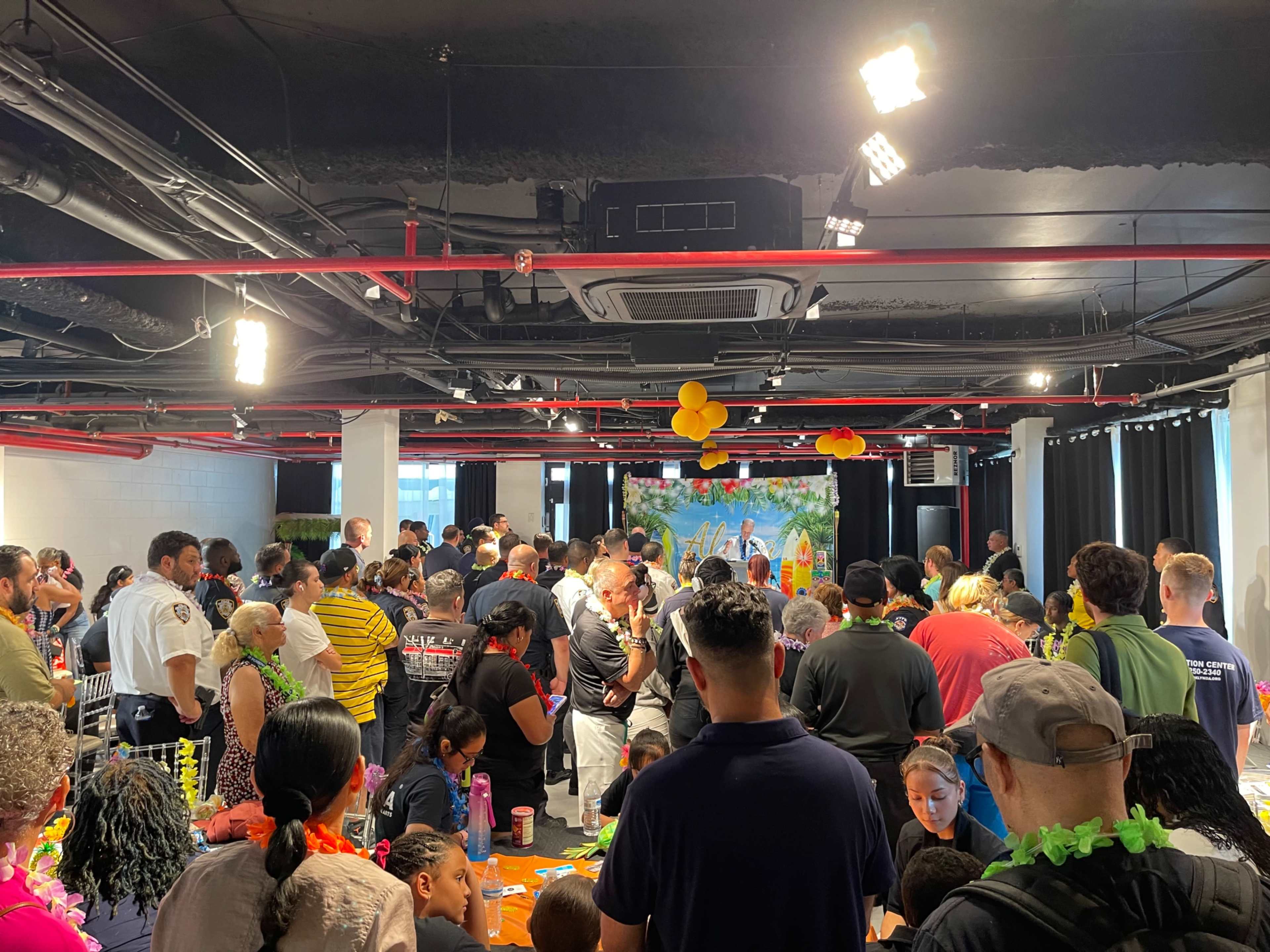 A large crowd gathers in a brightly lit indoor space with a mural in the background while people engage in various activities at tables.
