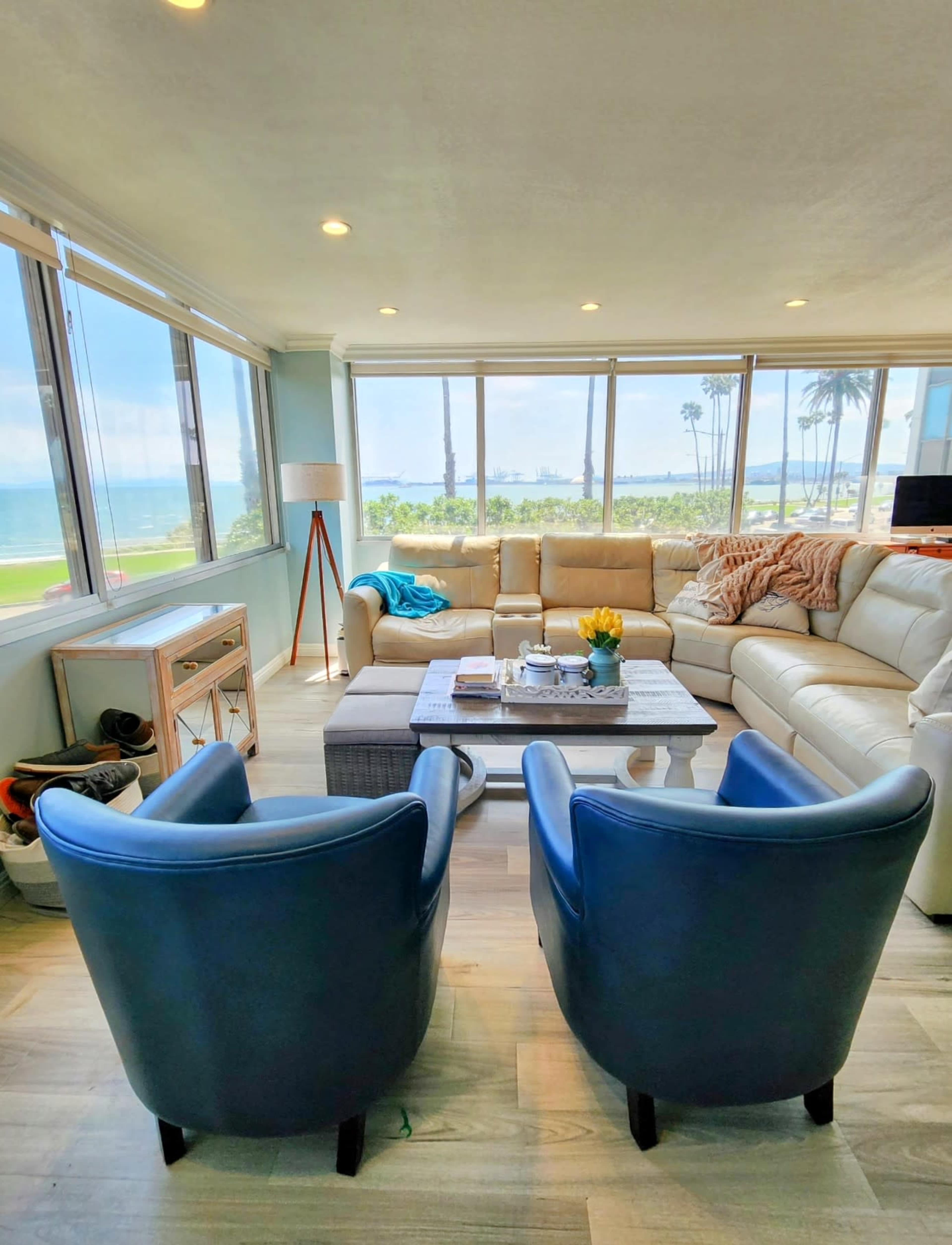 The scene shows a bright living room with a large sectional sofa and two blue chairs, overlooking a beach and palm trees through large windows.