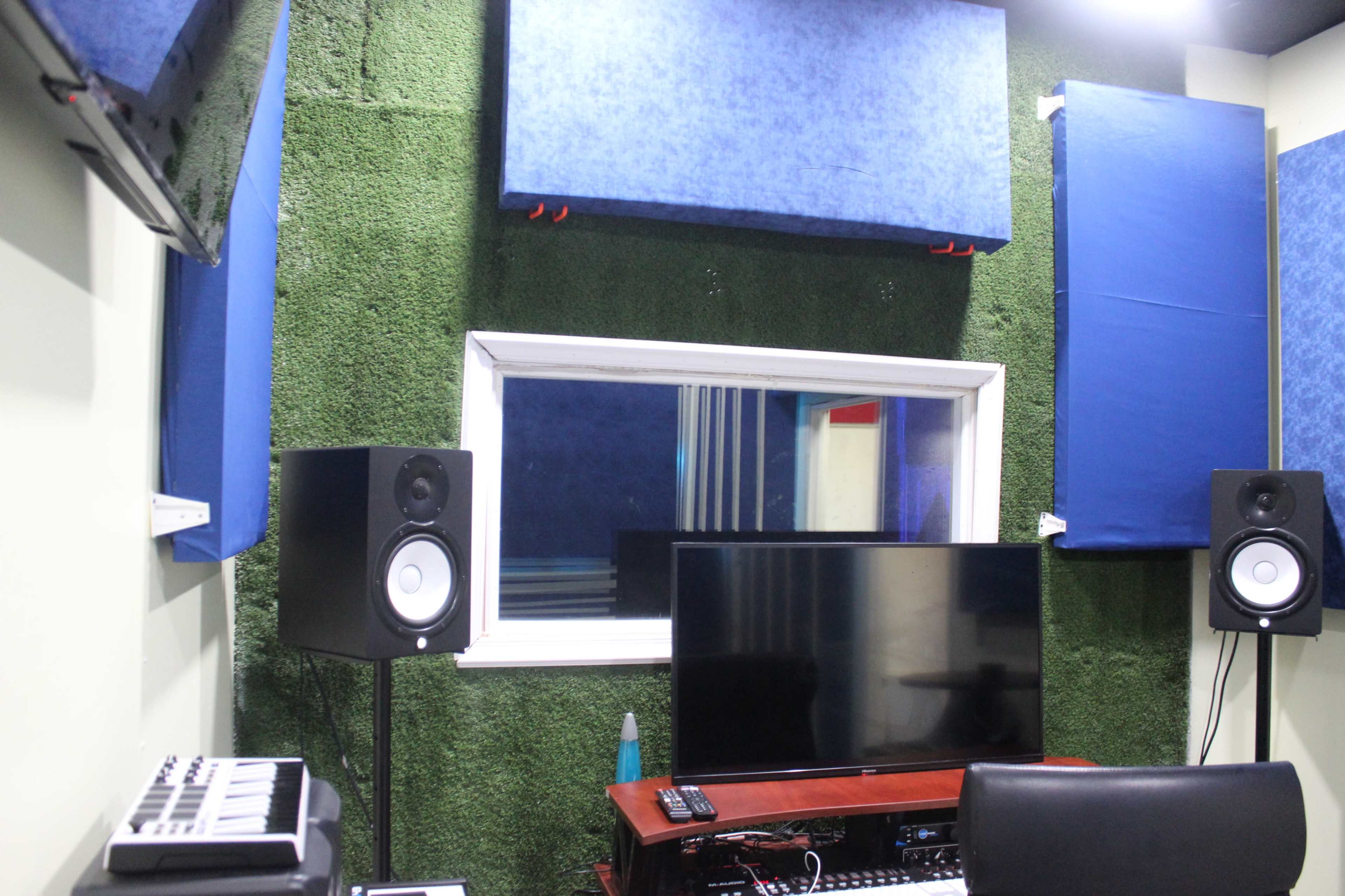 The image shows a sound studio with acoustic panels on the walls, a monitor on a desk, and studio speakers on either side of a window.