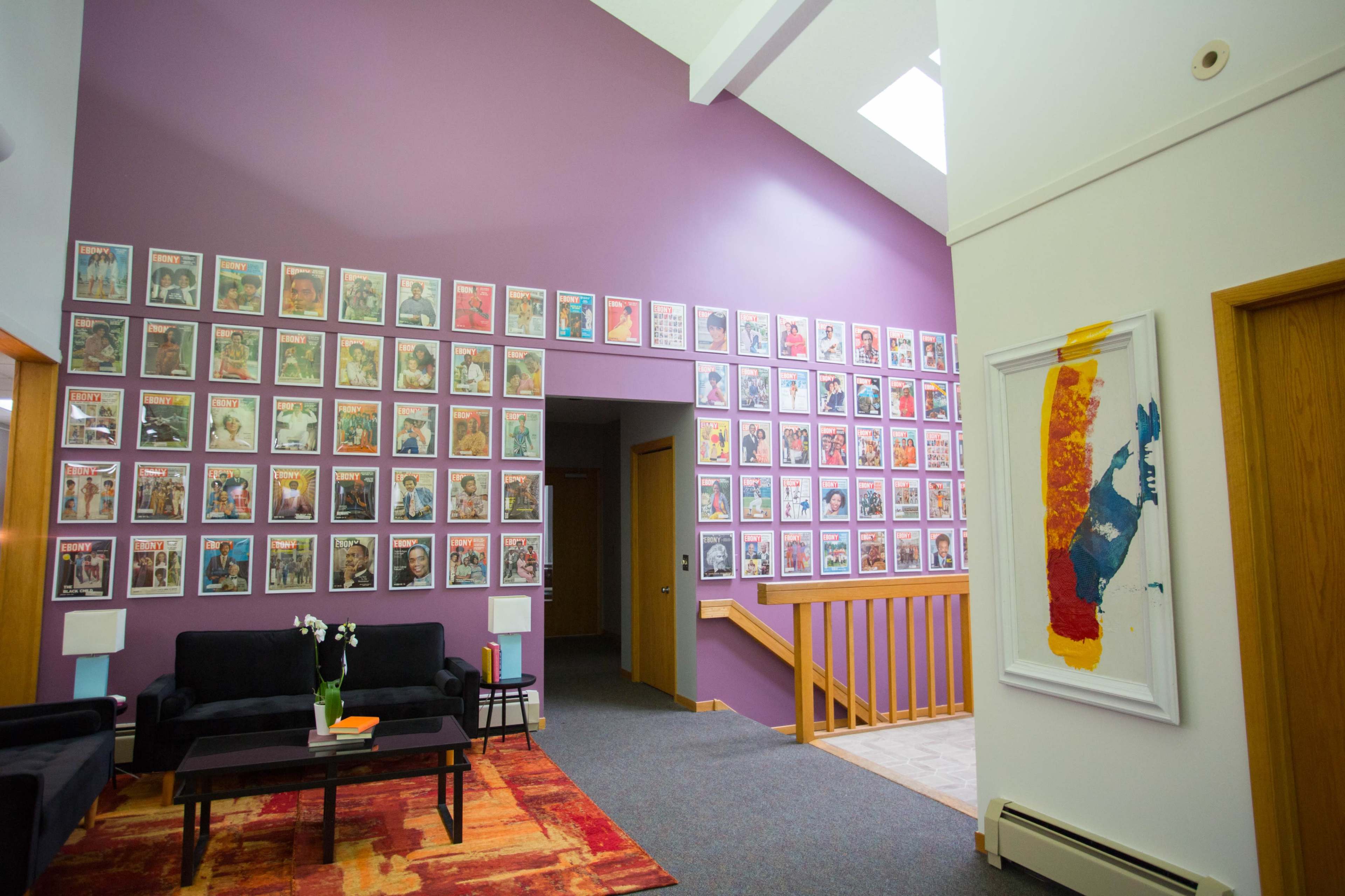 A brightly lit interior space featuring a wall adorned with numerous framed artworks in vibrant colors, complemented by a black sofa and wooden staircase.
