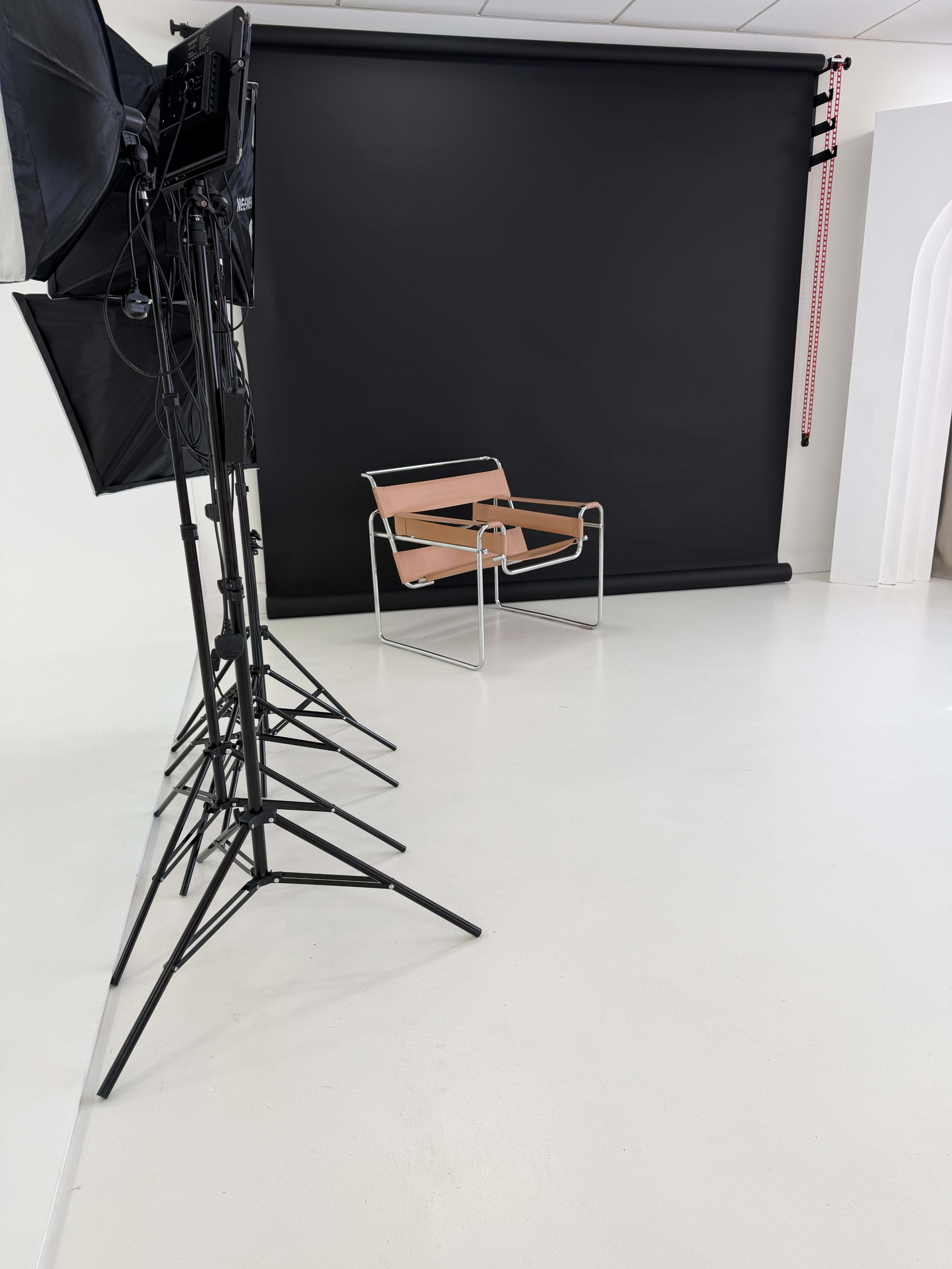 The image shows a minimalist studio setup with a black backdrop, a single modern chair, and light stands arranged in a corner.