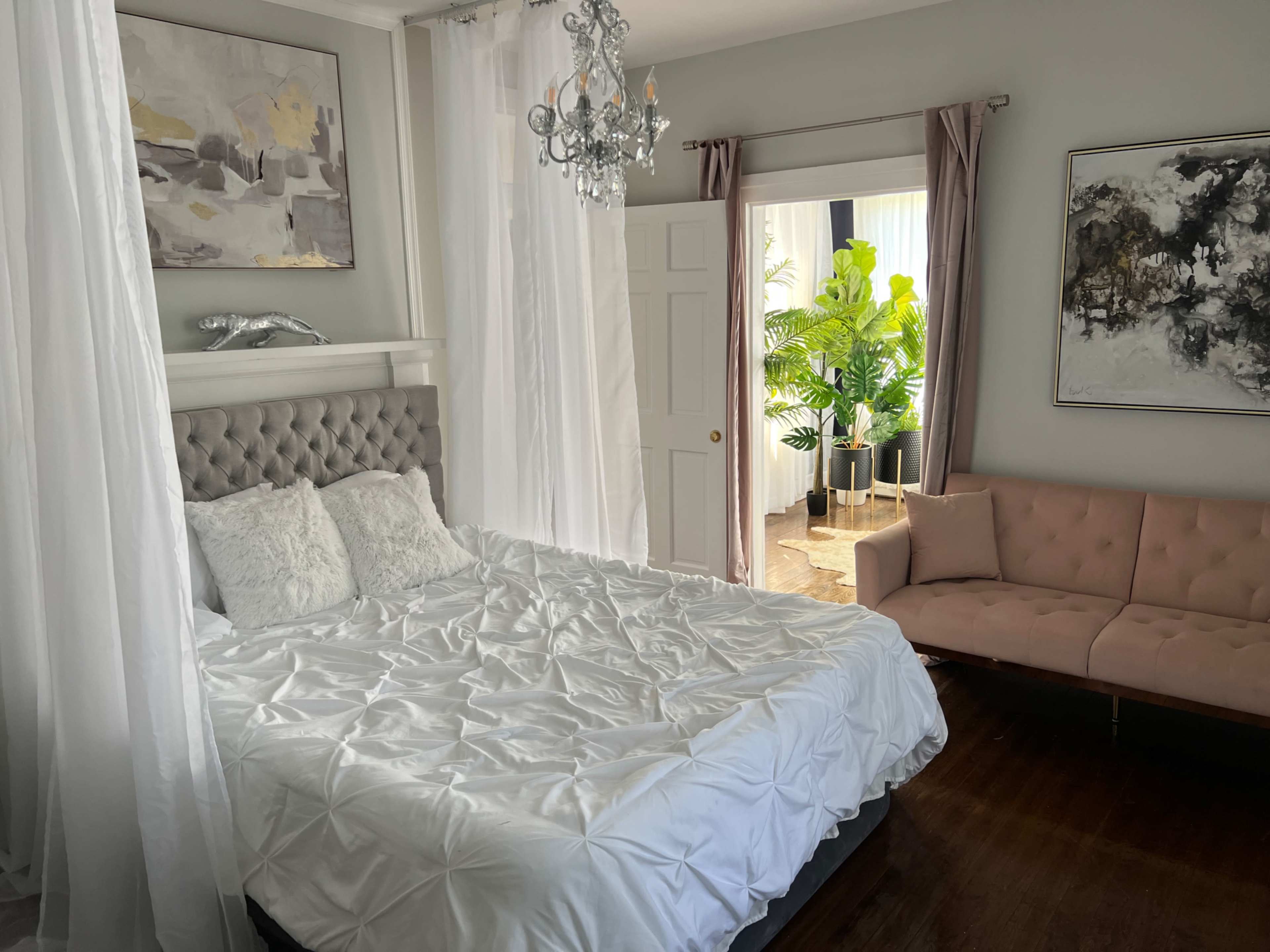 A neatly arranged bedroom features a tufted bed with a white duvet, a pink sofa, and a doorway leading to a plant-filled room.