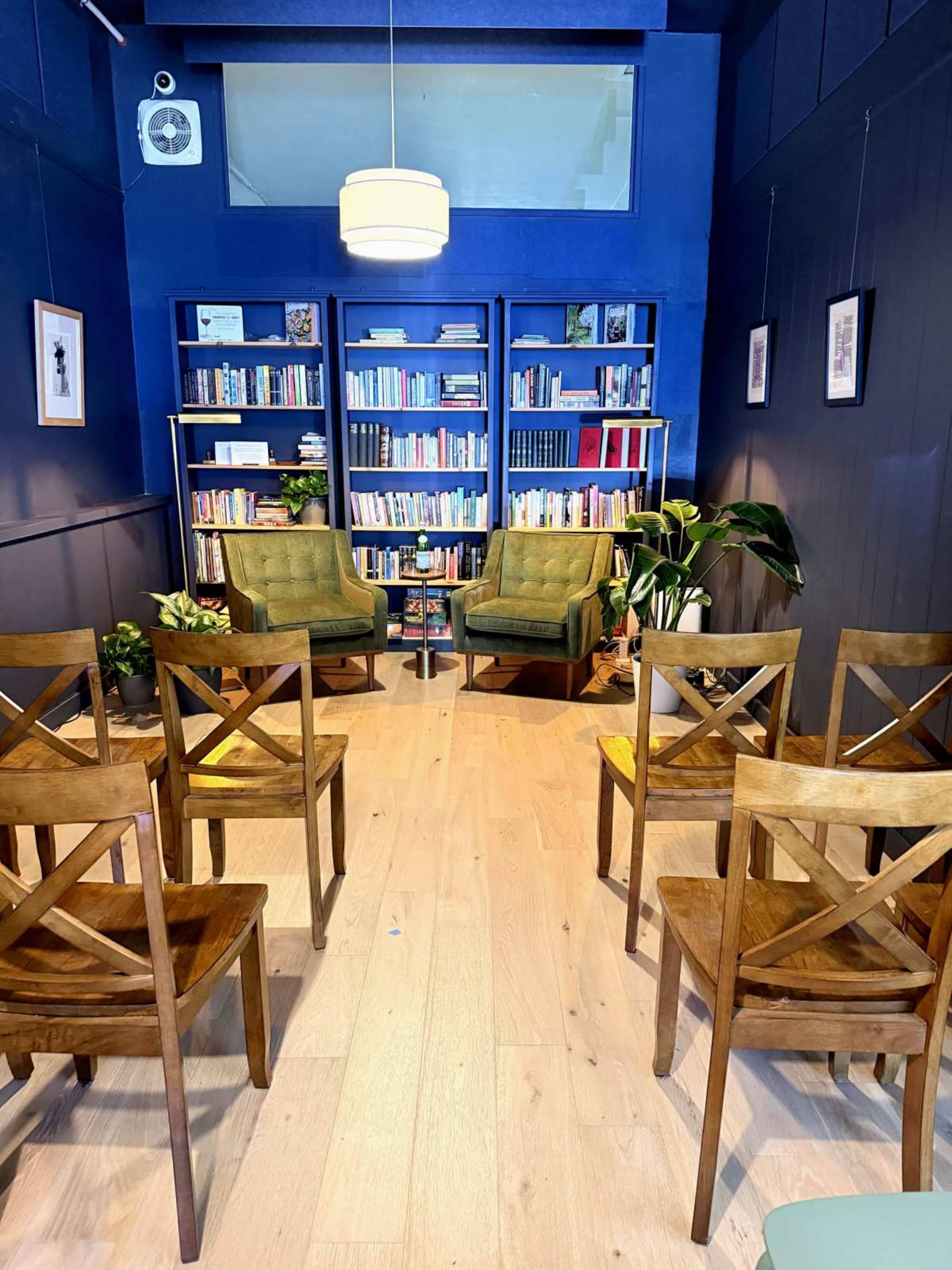 The image shows a cozy reading nook with two green armchairs facing a bookshelf, surrounded by wooden chairs and indoor plants.
