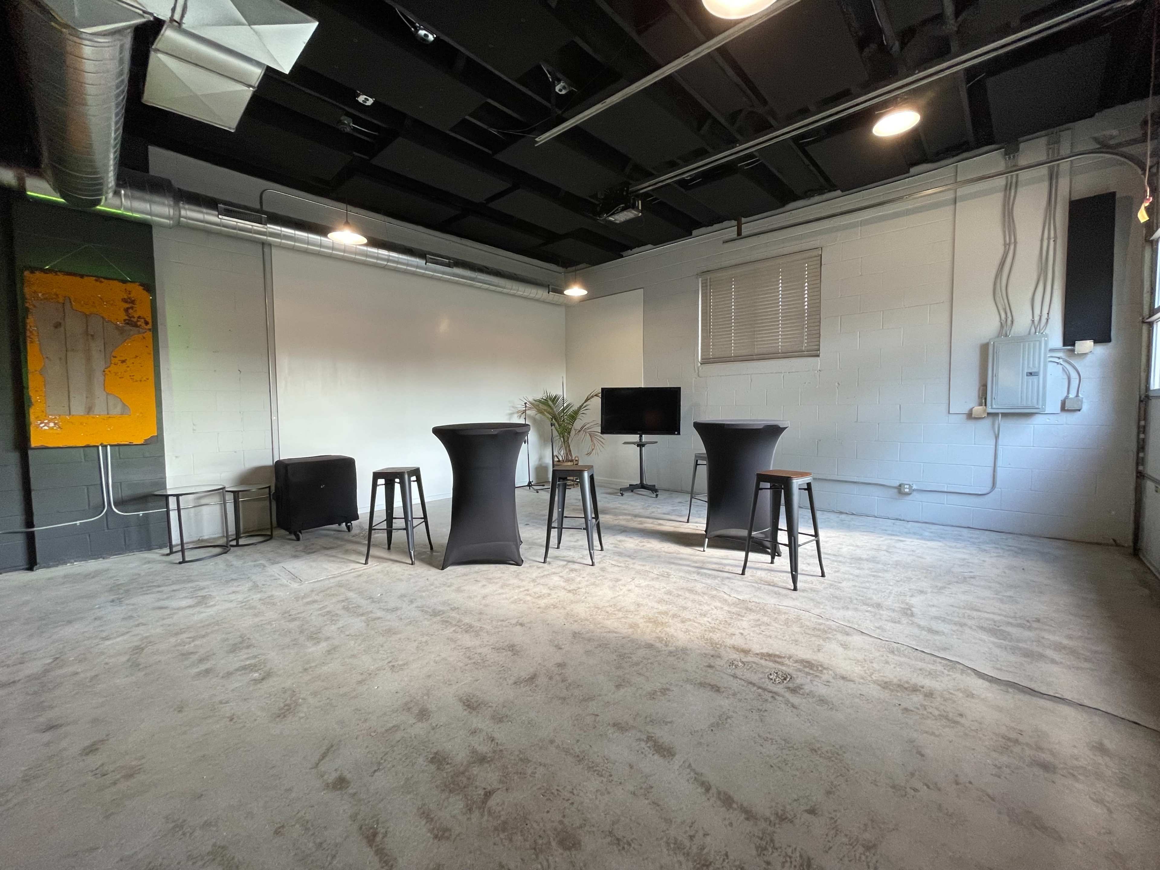 A sparse room with a concrete floor, featuring two tall black tables, a television on a stand, and a leafy plant near the back wall.