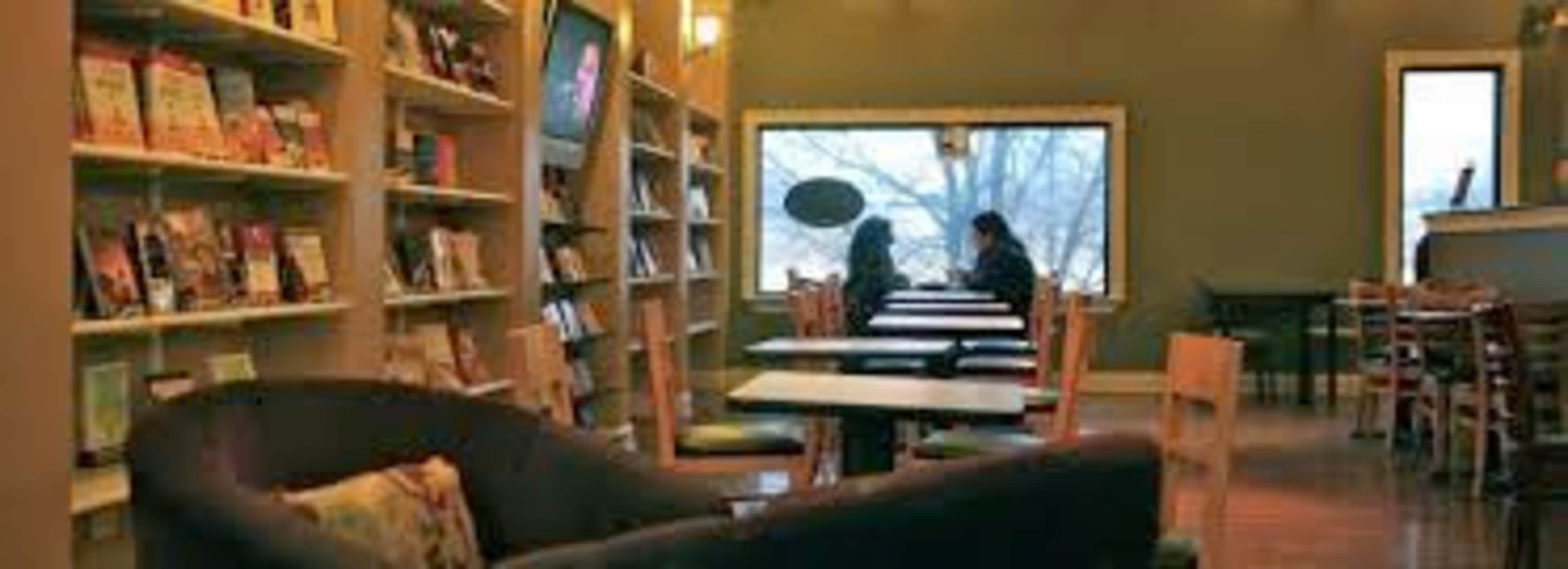 A cozy café features bookshelves lining the walls, empty tables, and two women seated by a window.