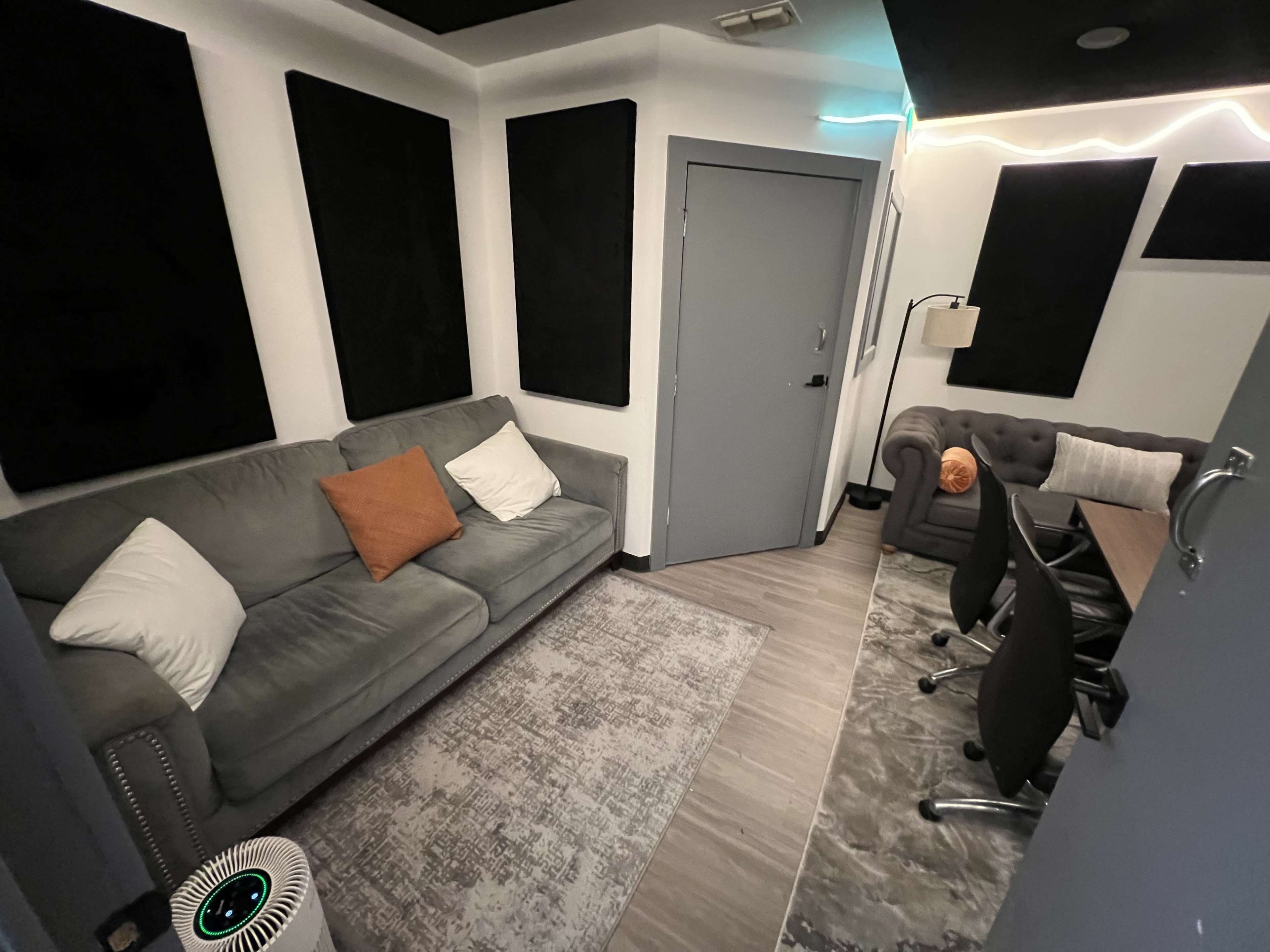 A small, soundproof room with a gray sofa, a chairs, and acoustic panels on the walls.