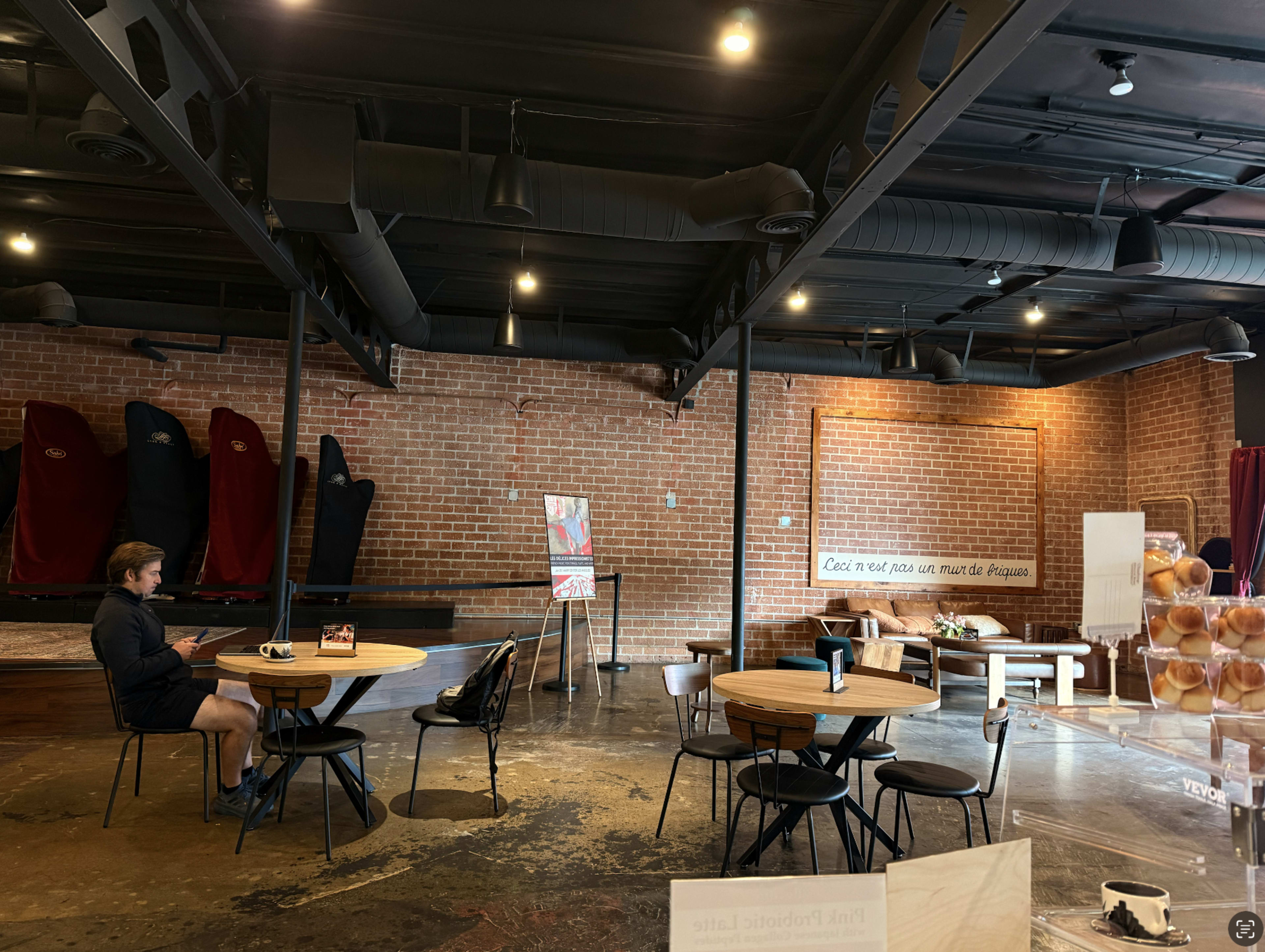The image shows a spacious café interior with exposed brick walls, wooden tables and chairs, and a seating area featuring a sofa against the wall.