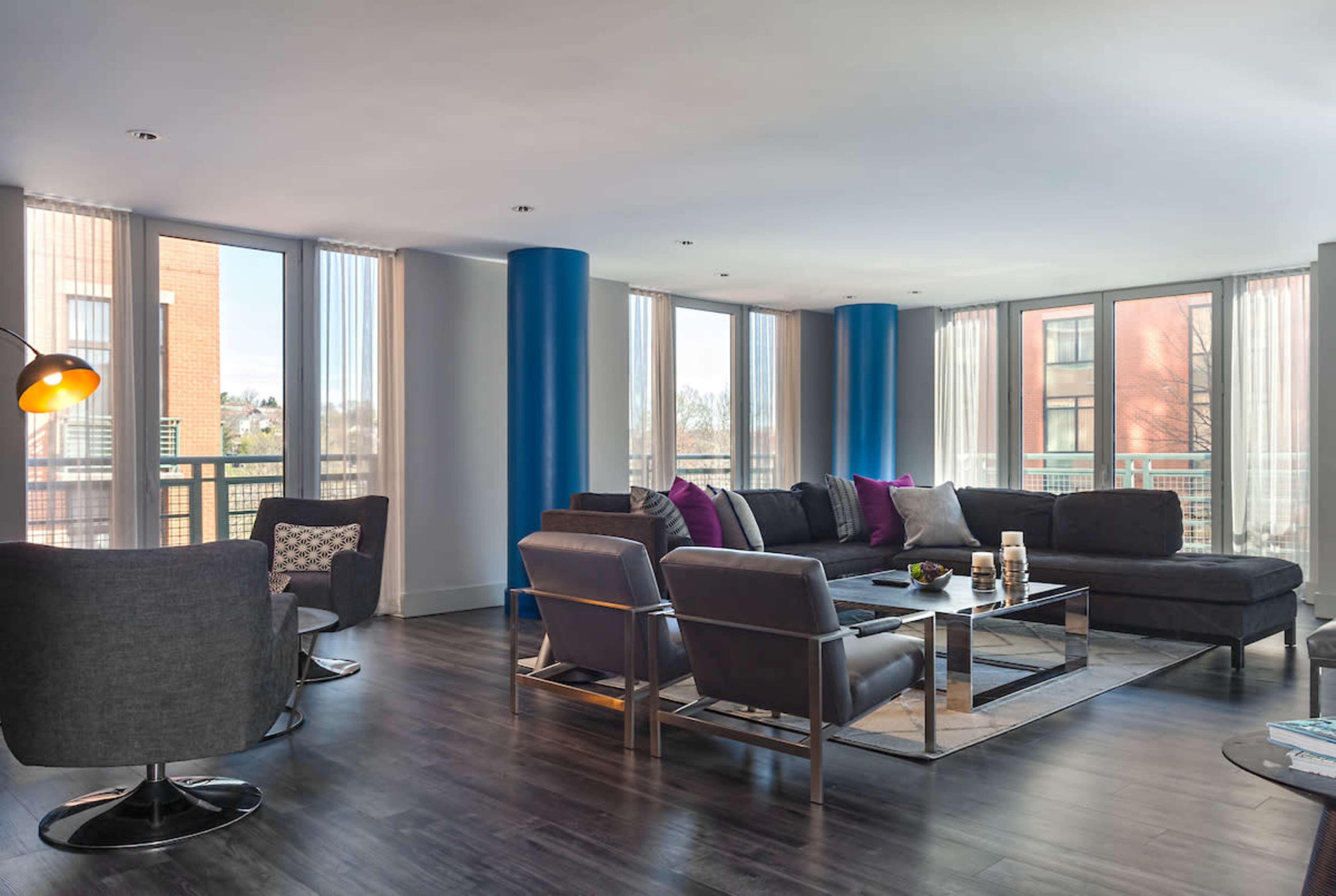 The image shows a modern living room with large windows, gray furniture, and vibrant accents.