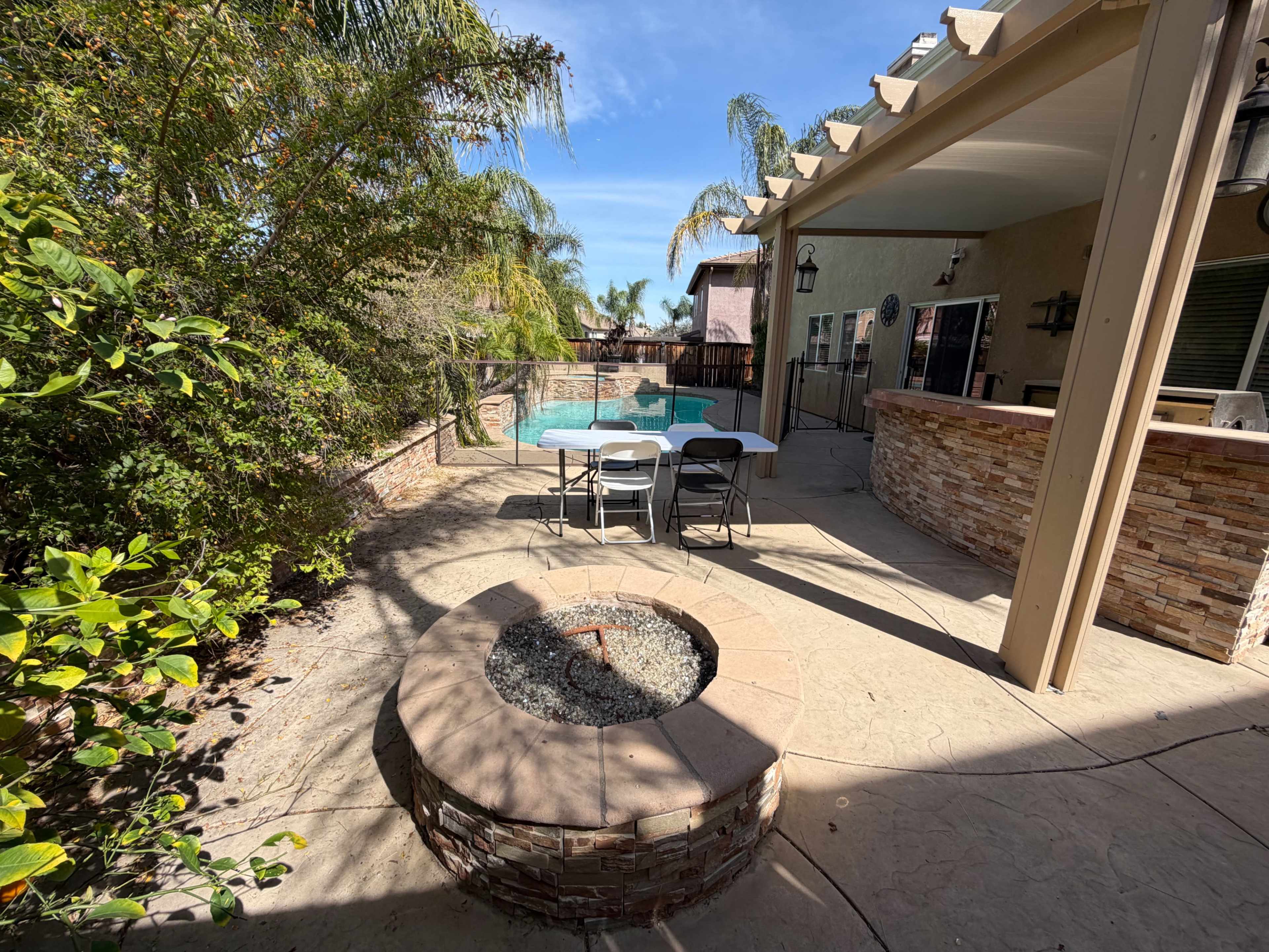 The image shows a backyard with a stone fire pit, a table and chairs, and a swimming pool surrounded by tropical plants.
