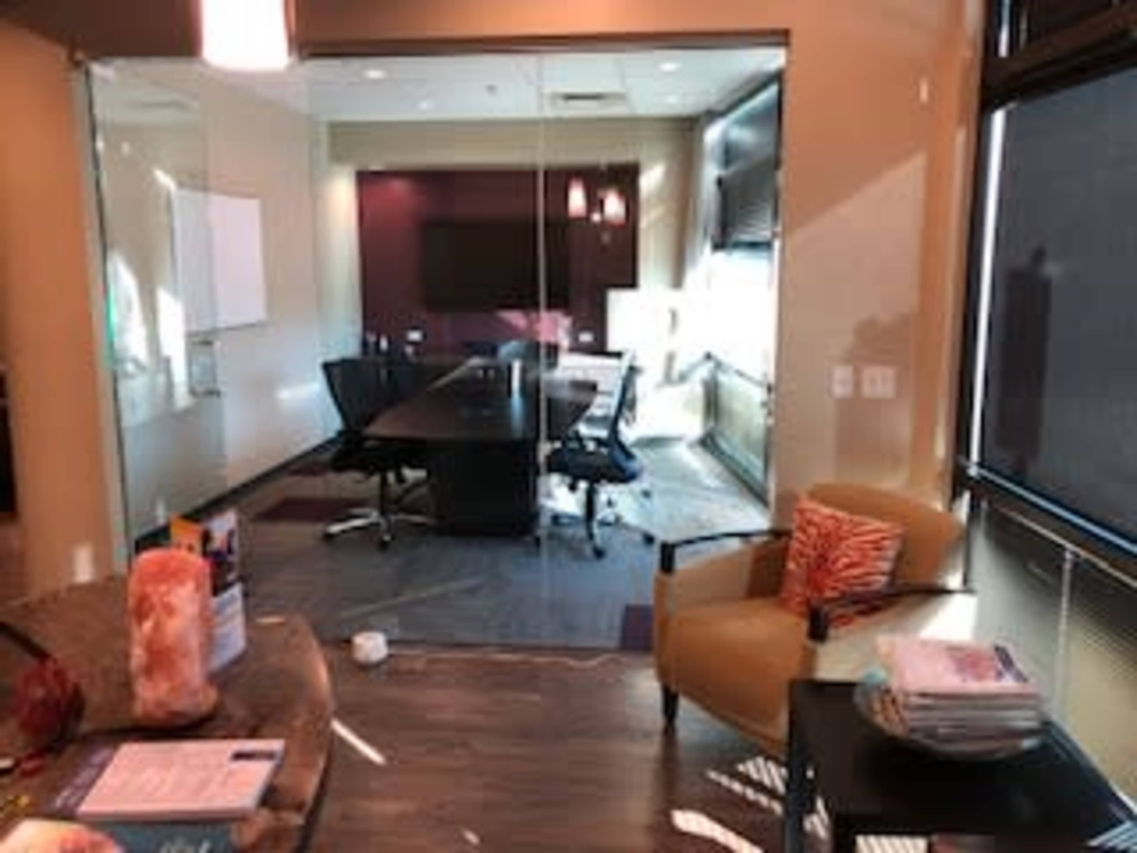 A modern conference room with a large table, black chairs, and a wall-mounted screen, featuring a partially glass wall and warm lighting.