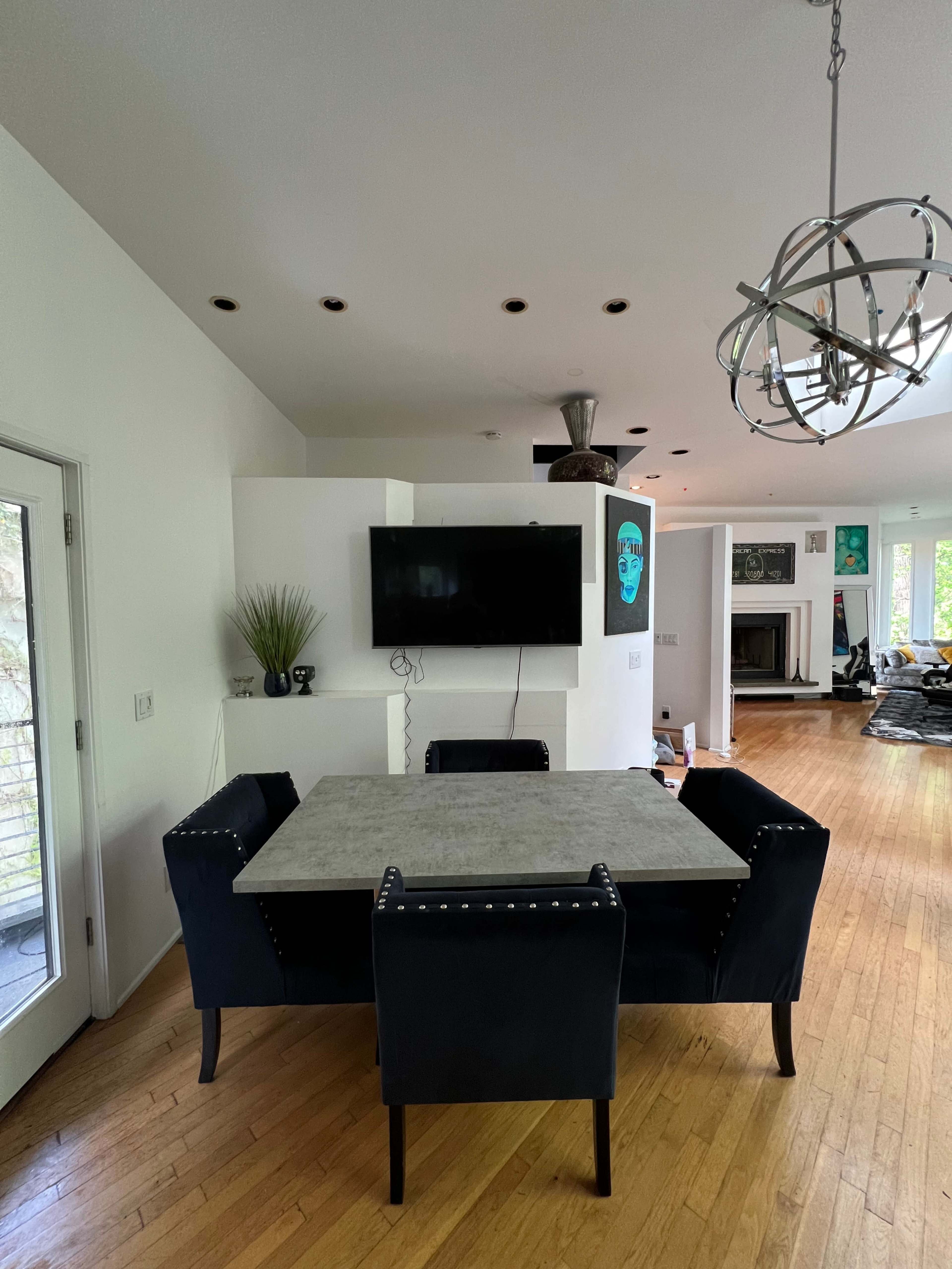 A dining area features a rectangular table surrounded by four upholstered chairs, with a wall-mounted television and an overhead light fixture above.