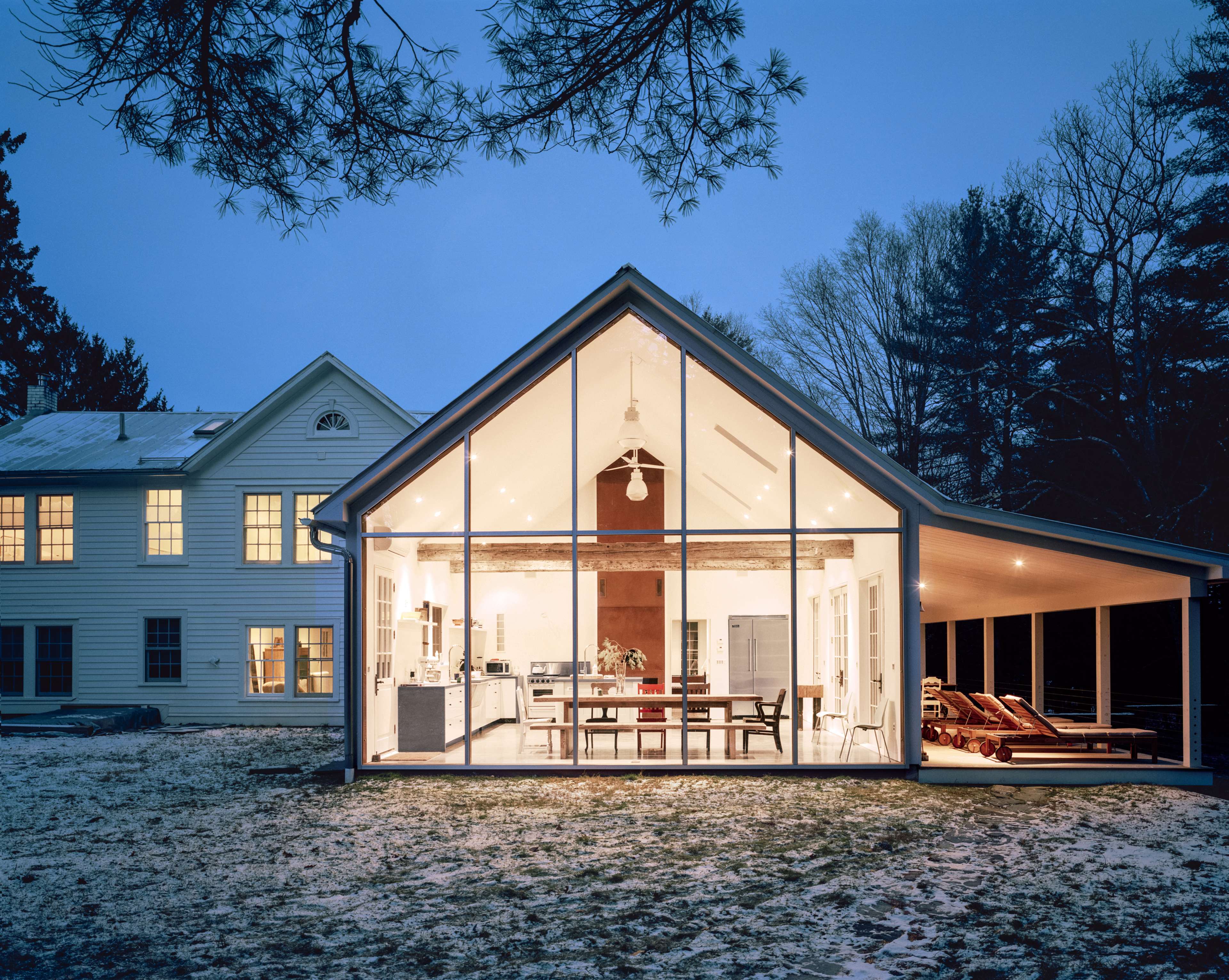 A modern, glass-fronted extension of a house is illuminated at dusk, showcasing a spacious interior with a dining area and kitchen.