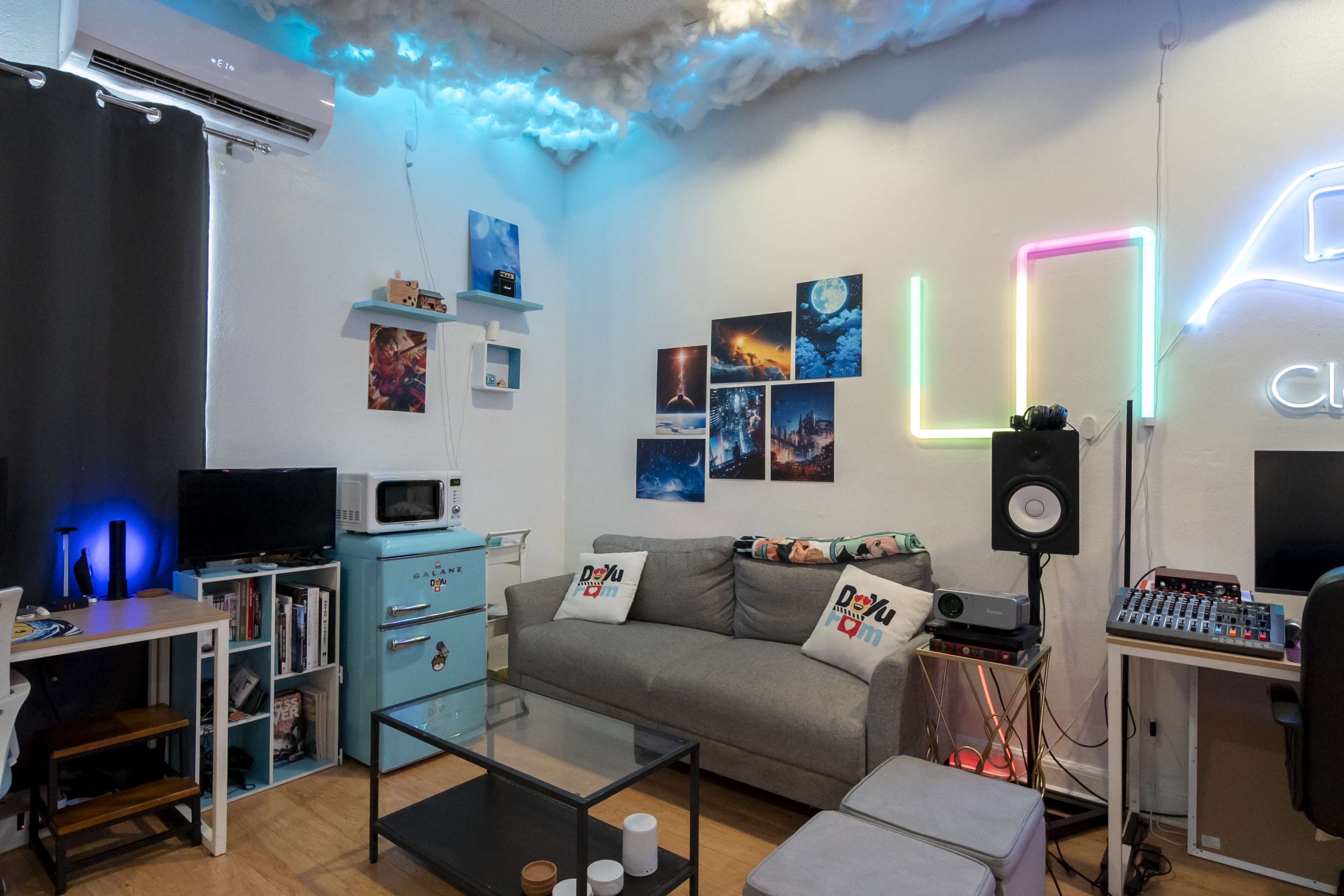 The image shows a cozy living space featuring a gray sofa, a coffee table, and a wall decorated with various framed pictures and colorful lights.