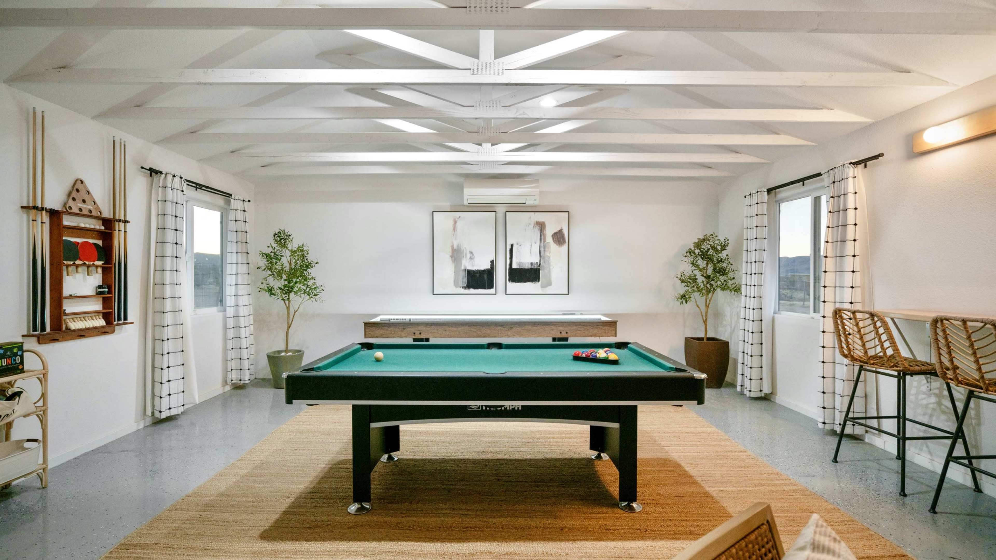 The image displays a spacious game room featuring a green pool table, a wooden game table, and two large windows with curtains.