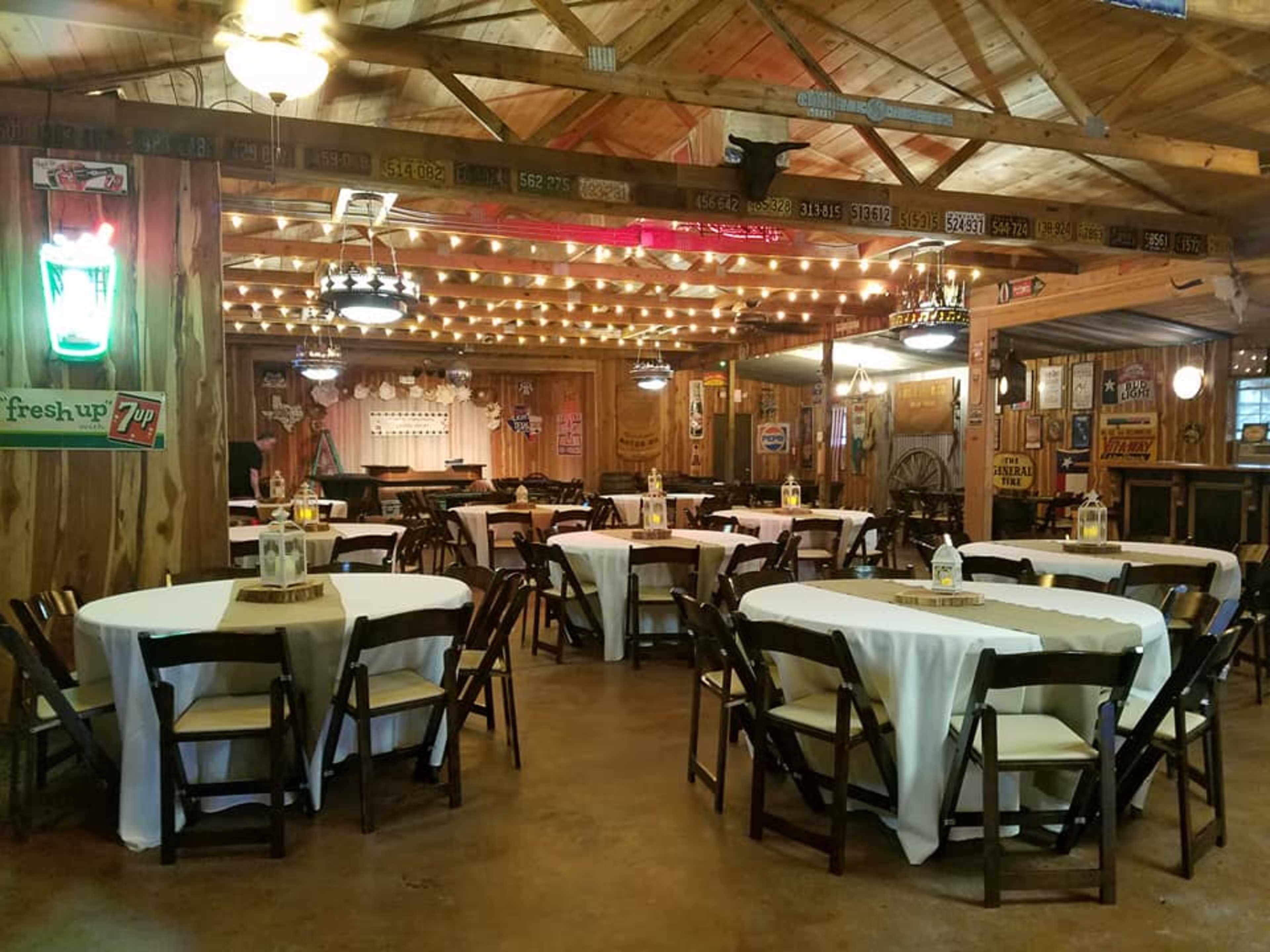 A rustic indoor event space with wooden walls, round tables covered with white tablecloths, and string lights hanging from the ceiling.