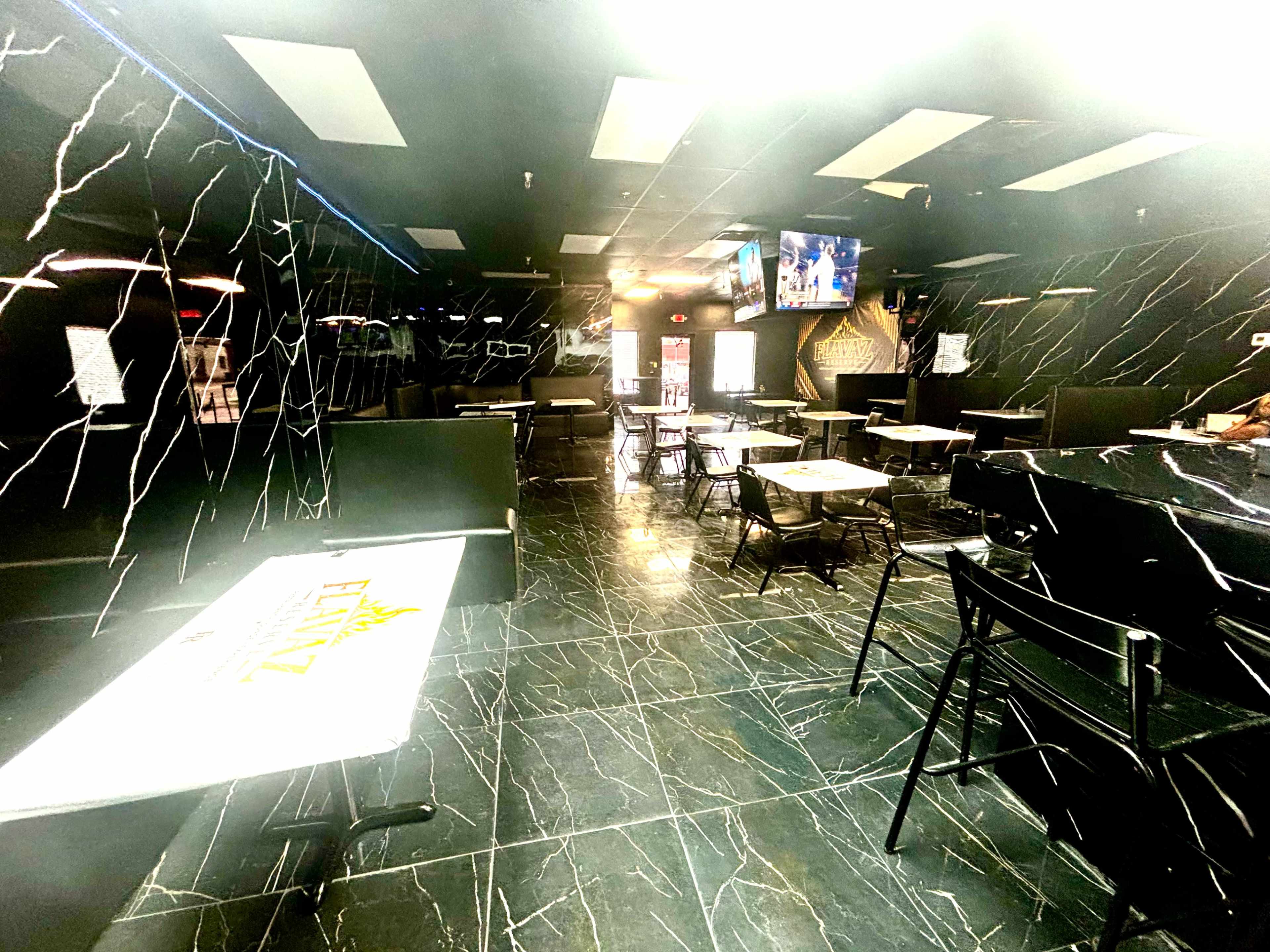 The image shows an empty restaurant with black marble flooring, dark walls, and tables set with chairs, along with a television screen mounted on the wall.