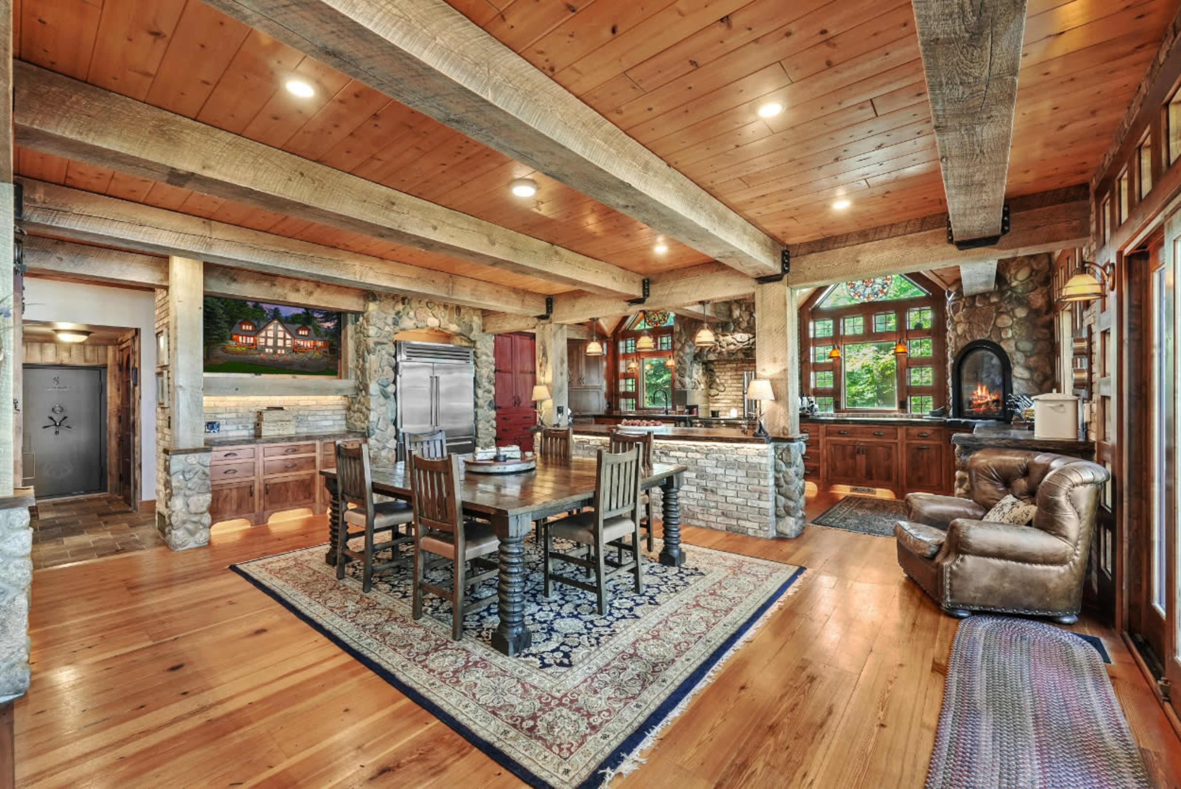 The image depicts a spacious kitchen and dining area with wooden beams, stone accents, and a large wooden table surrounded by chairs.