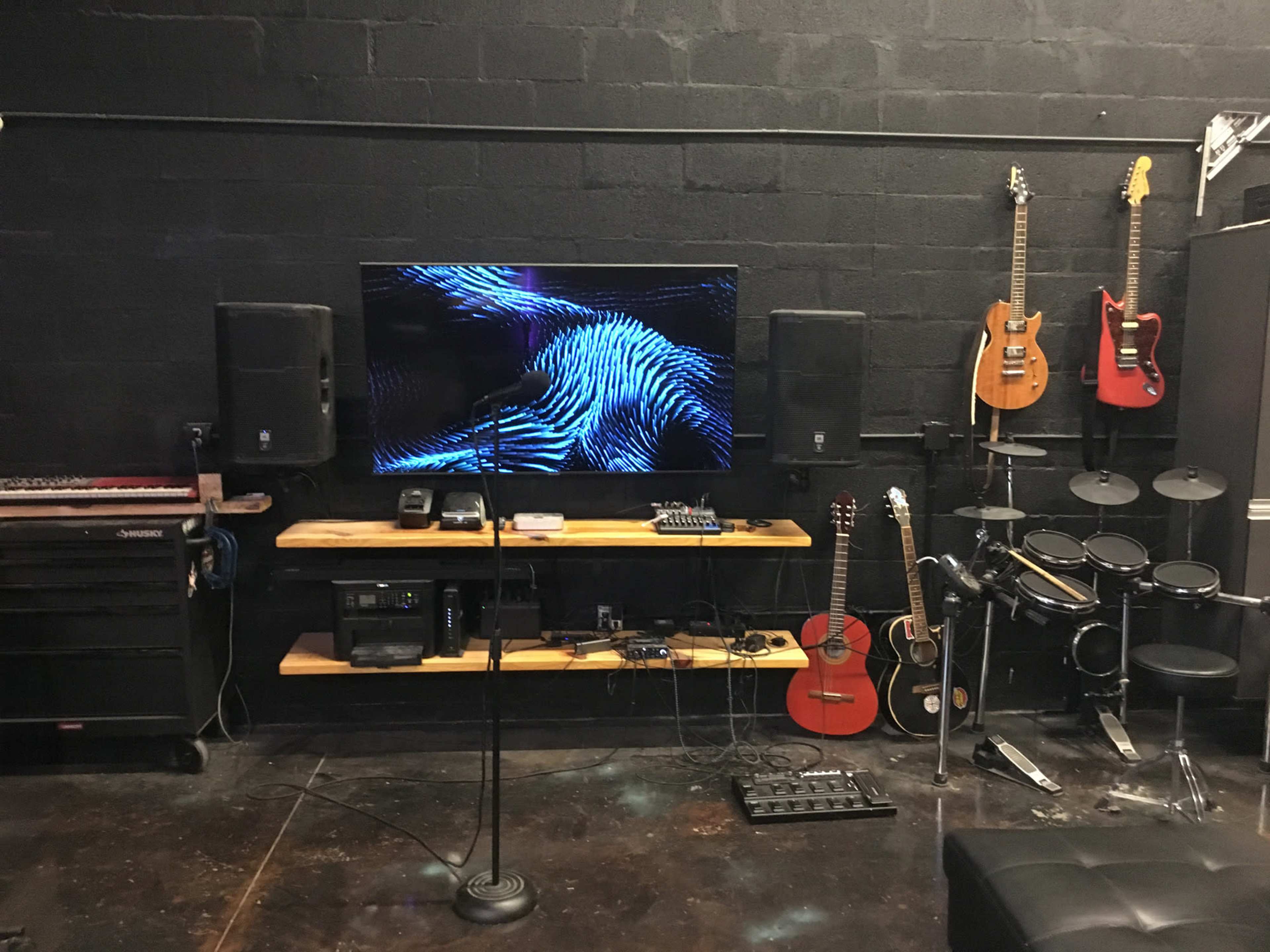 The image shows a music studio setup with a large television, speakers, a microphone stand, various musical instruments, and equipment along a wooden shelf against a textured black wall.