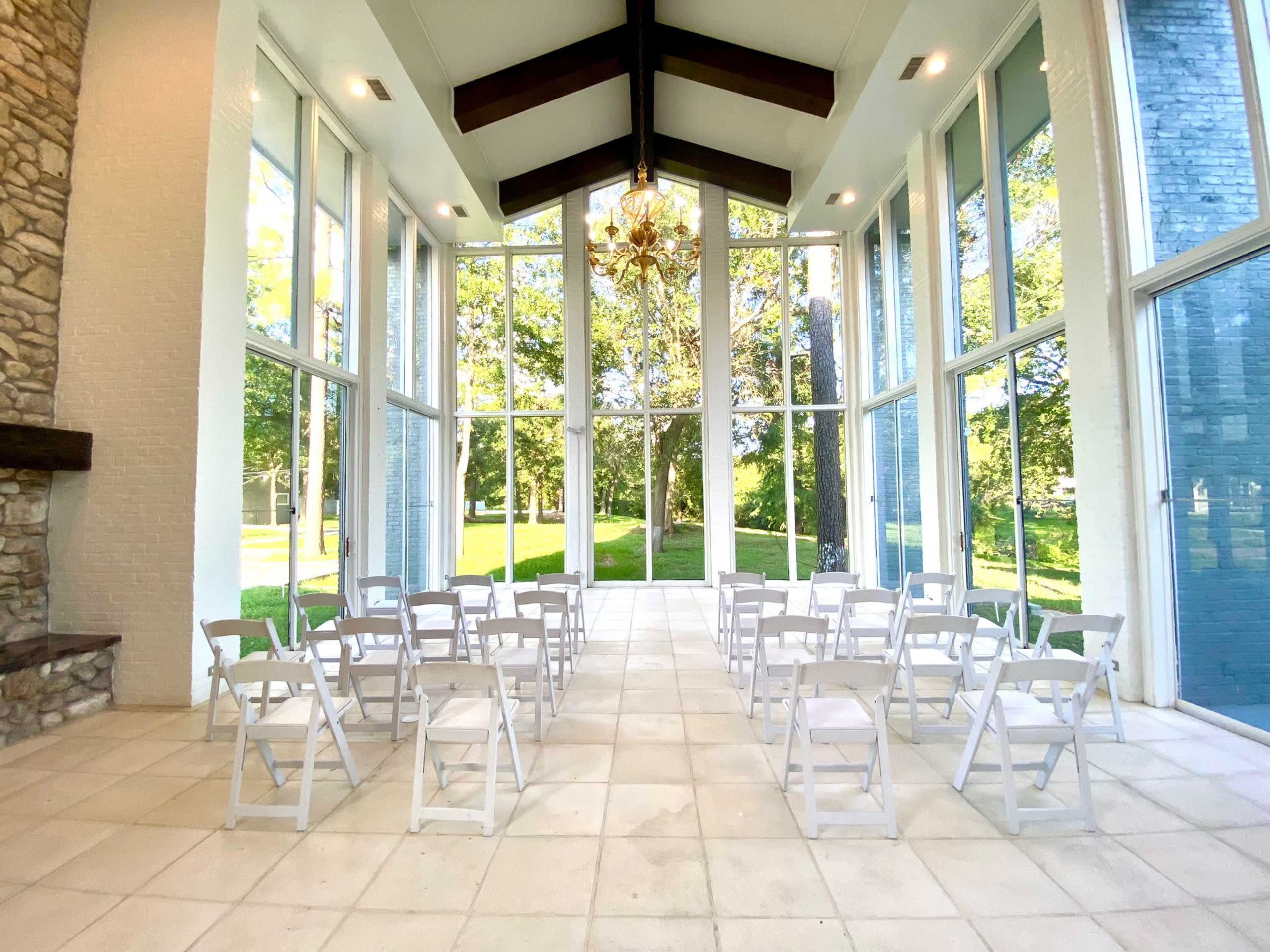 A spacious room with large windows overlooking a green landscape, arranged with white chairs in rows and a chandelier hanging from the ceiling.