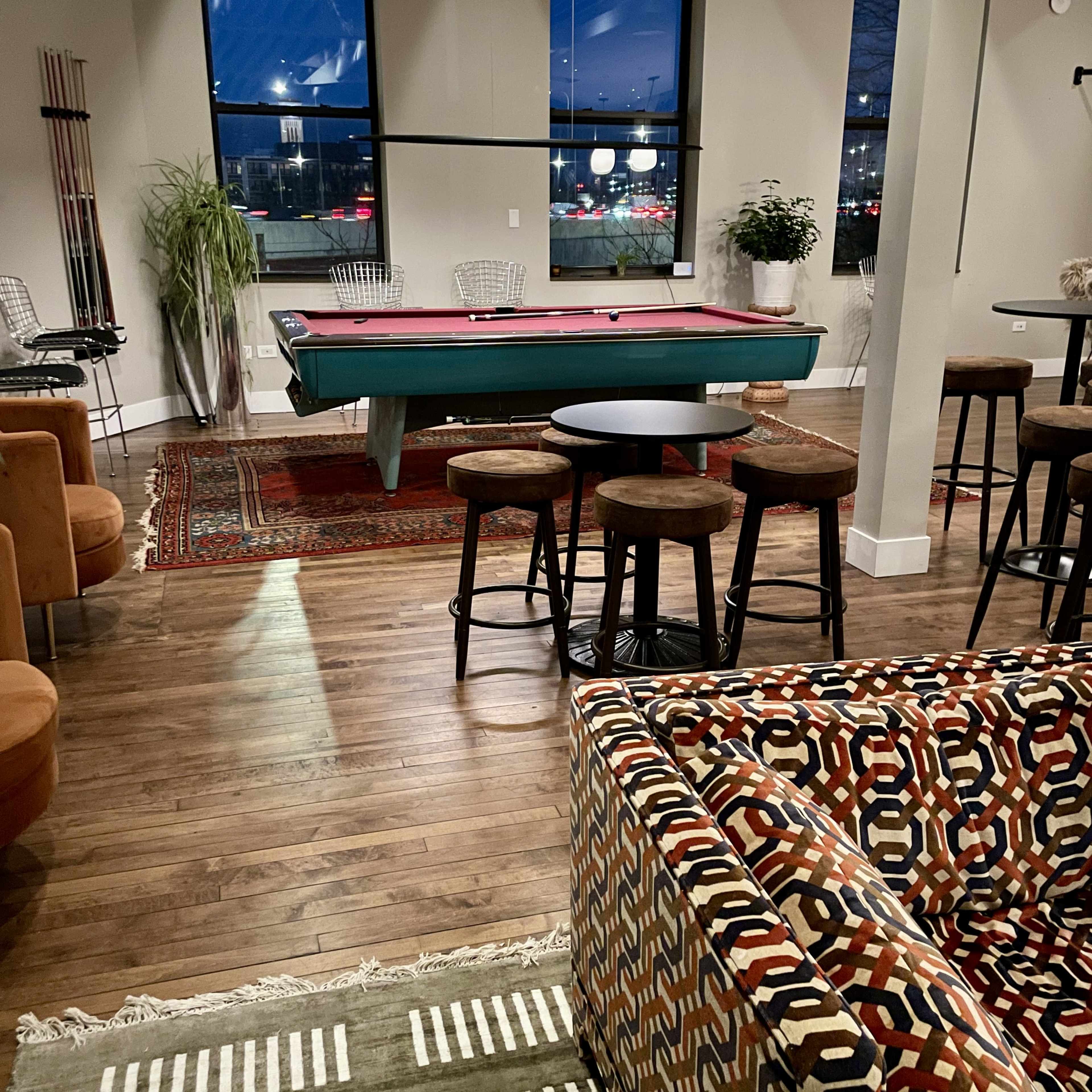 The image shows a modern lounge area featuring a pool table, several seating options, and large windows with a view of the outdoors.