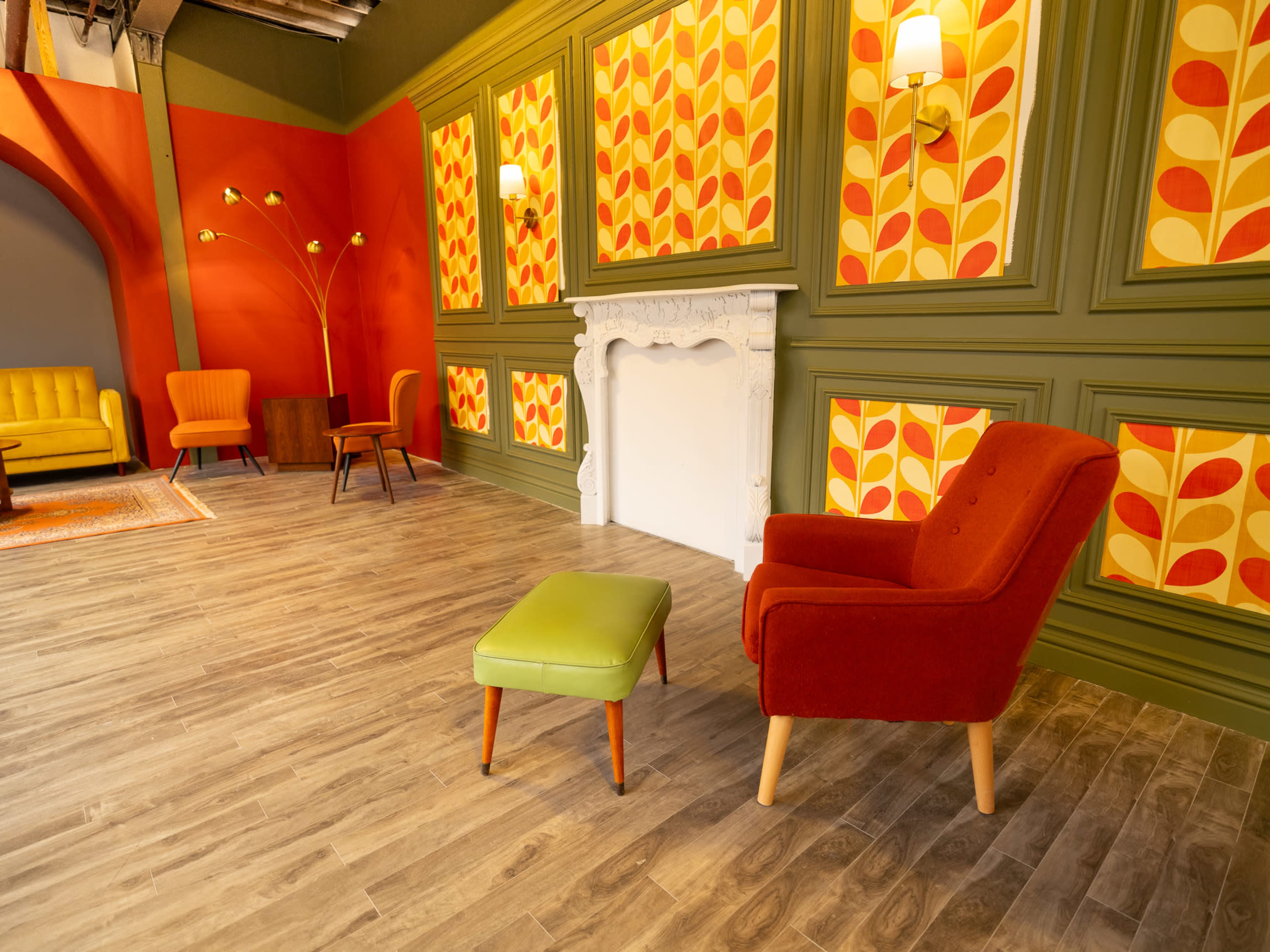 The image shows a stylized room with retro wallpaper featuring leaf patterns, a white fireplace, and a mix of colorful furniture including armchairs and an ottoman.