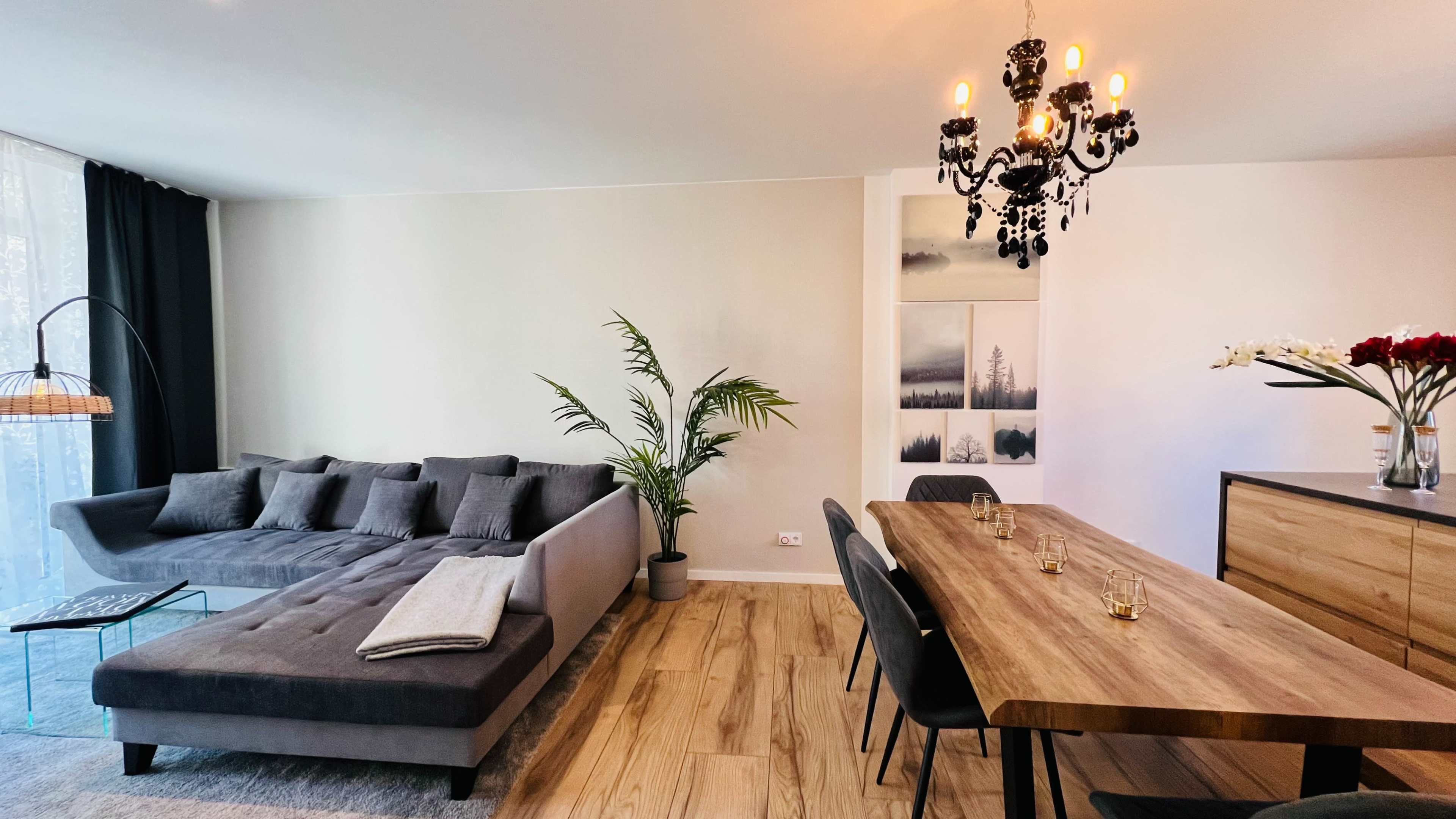 A modern living room with a gray sectional sofa, a wooden dining table set for several people, and decorative wall art alongside a plant in the corner.