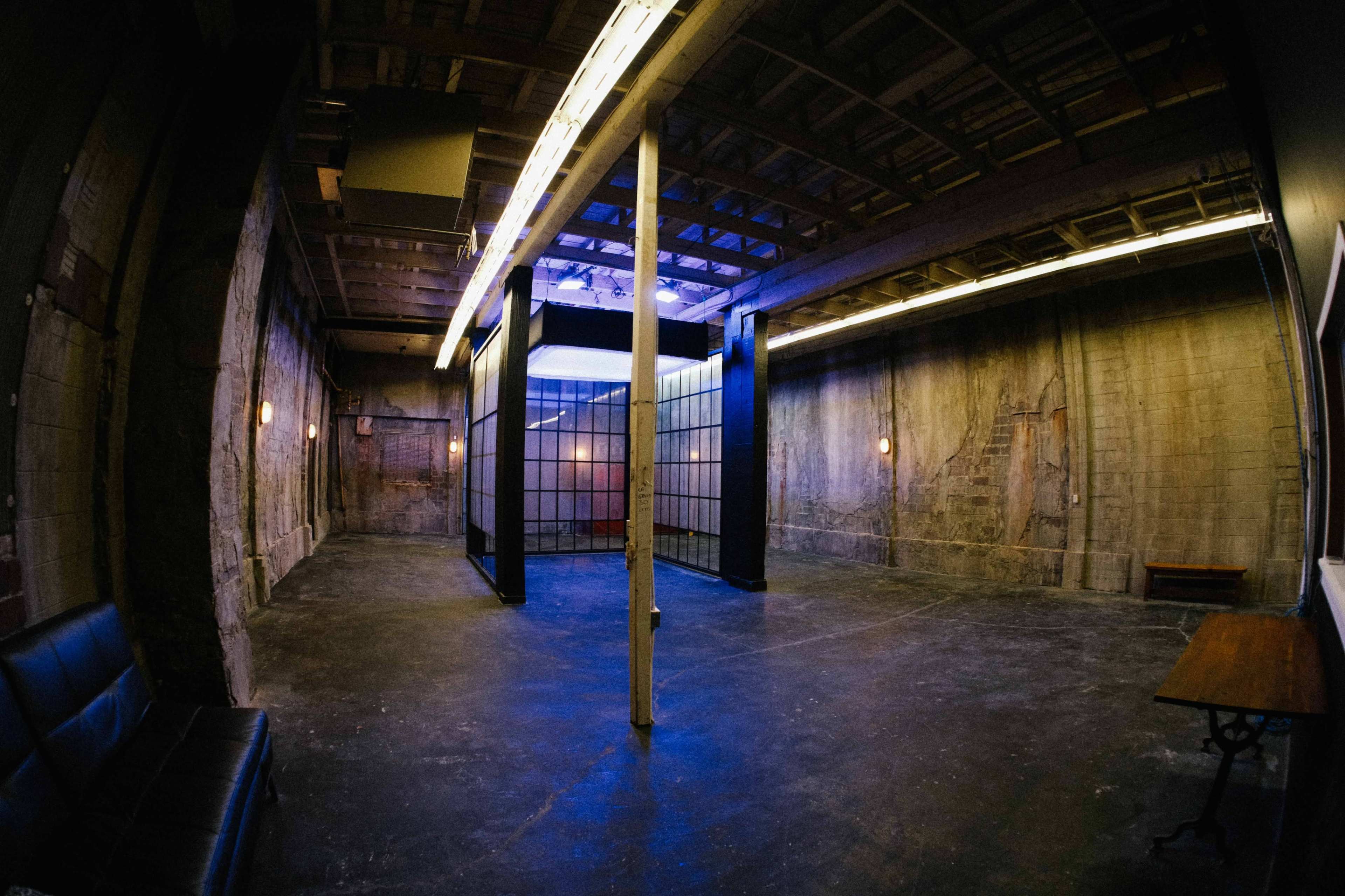 The image depicts an empty, industrial-style room with concrete walls, a grid partition, and sparse lighting.