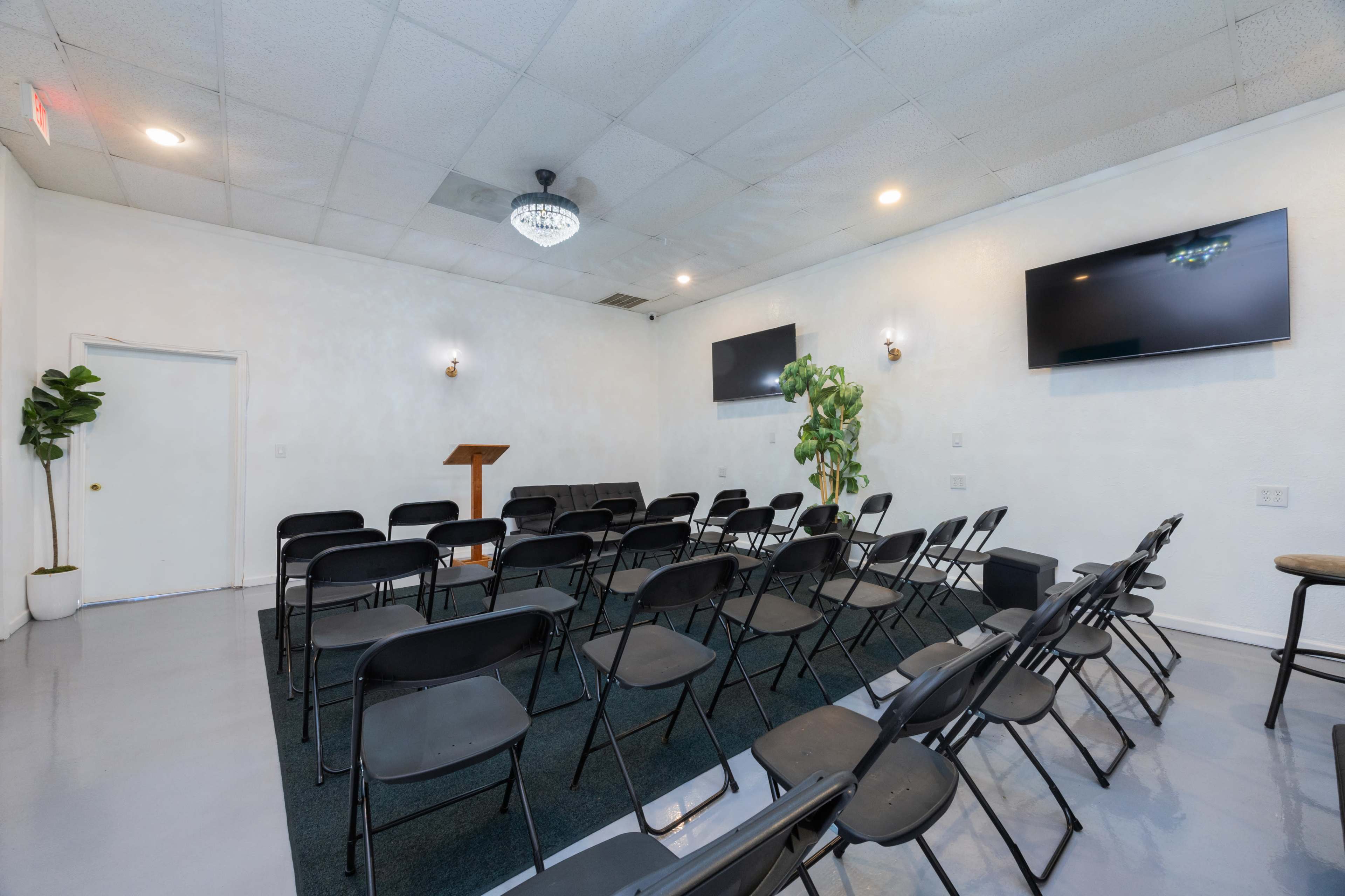 A small, empty room set up with rows of black chairs facing a wooden podium, flanked by two wall-mounted monitors and a potted plant in one corner.