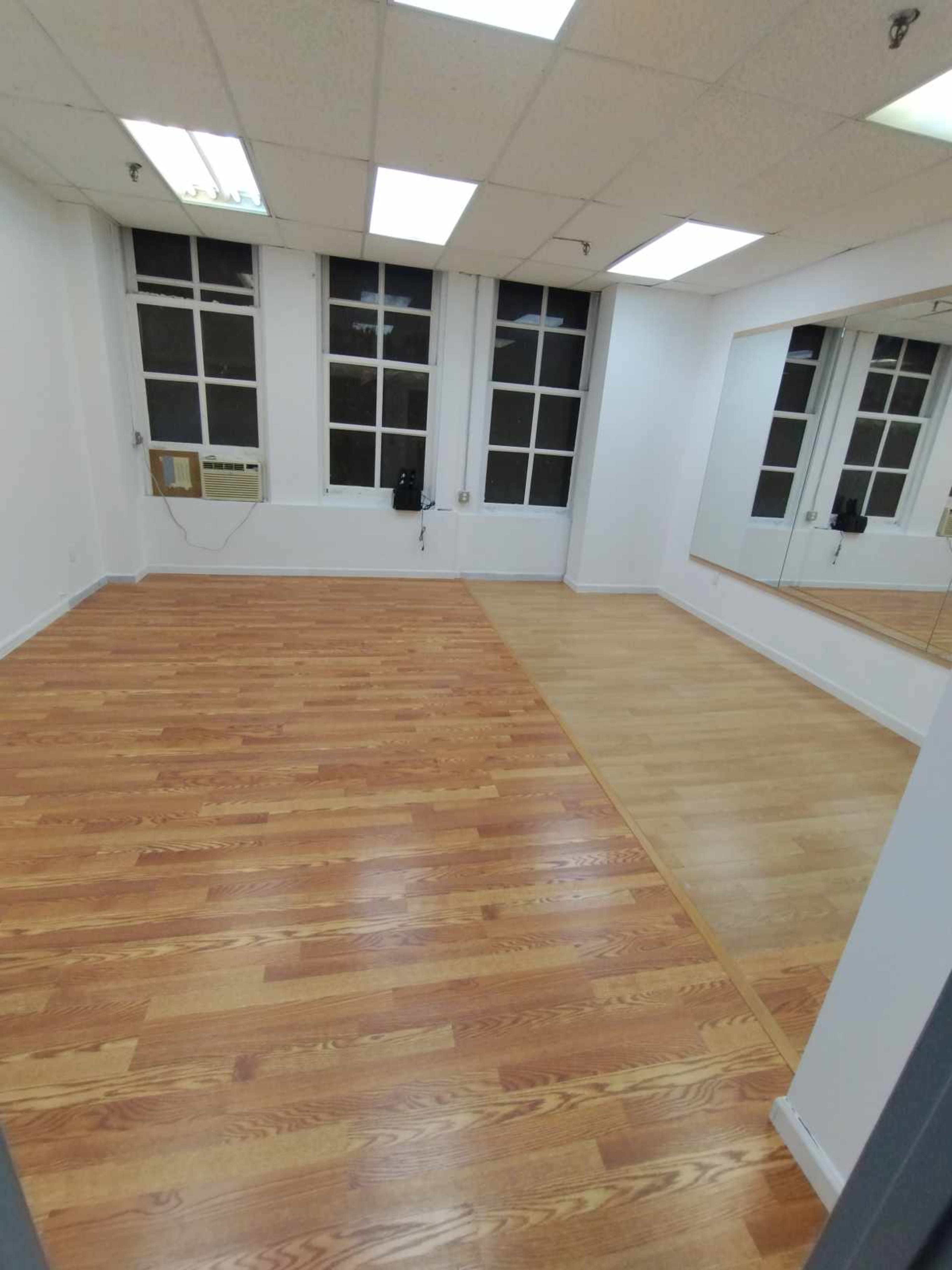 The image shows an empty, well-lit room with hardwood flooring and large windows, along with a mirror on one wall.