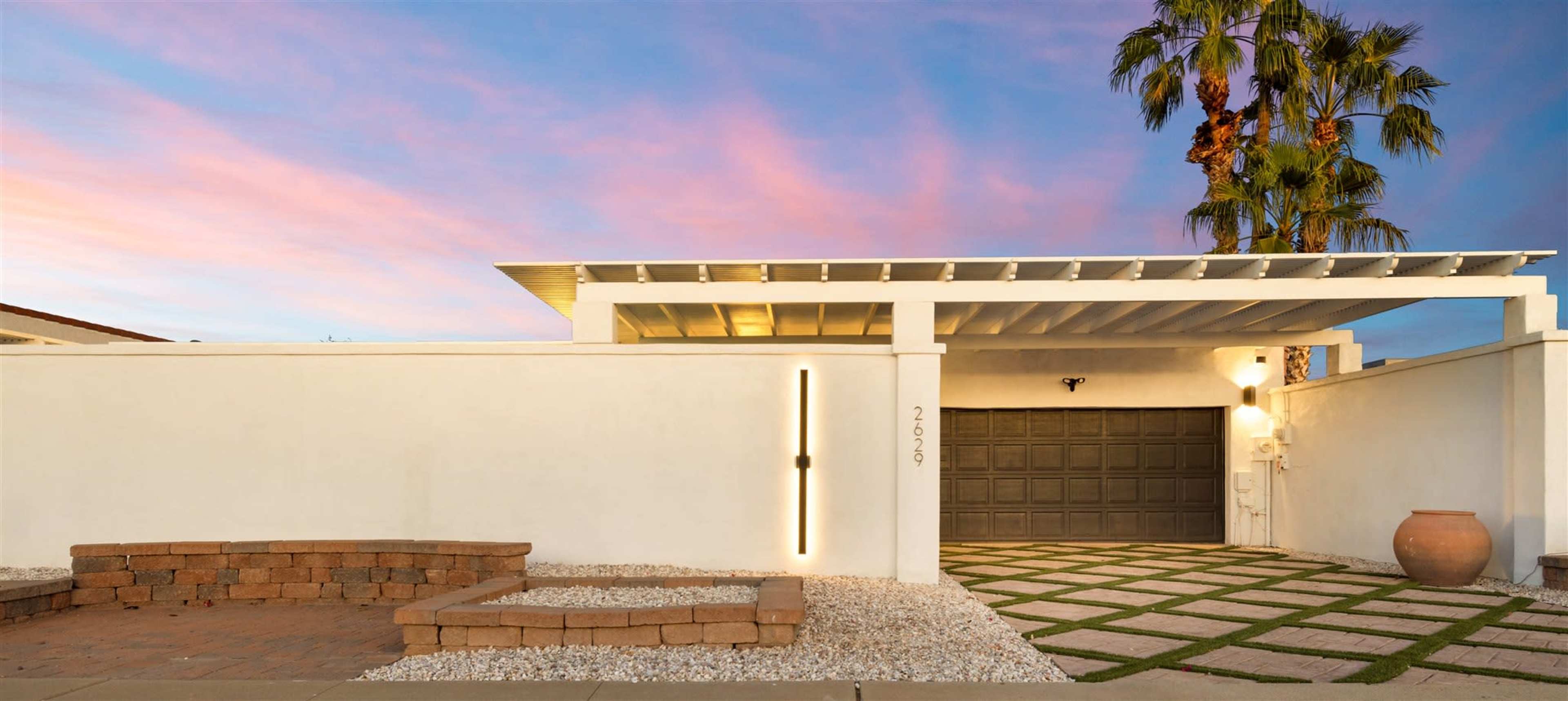 A modern home features a minimalist design with a gray garage door, a stone pathway, and a palm tree against a colorful sunset sky.