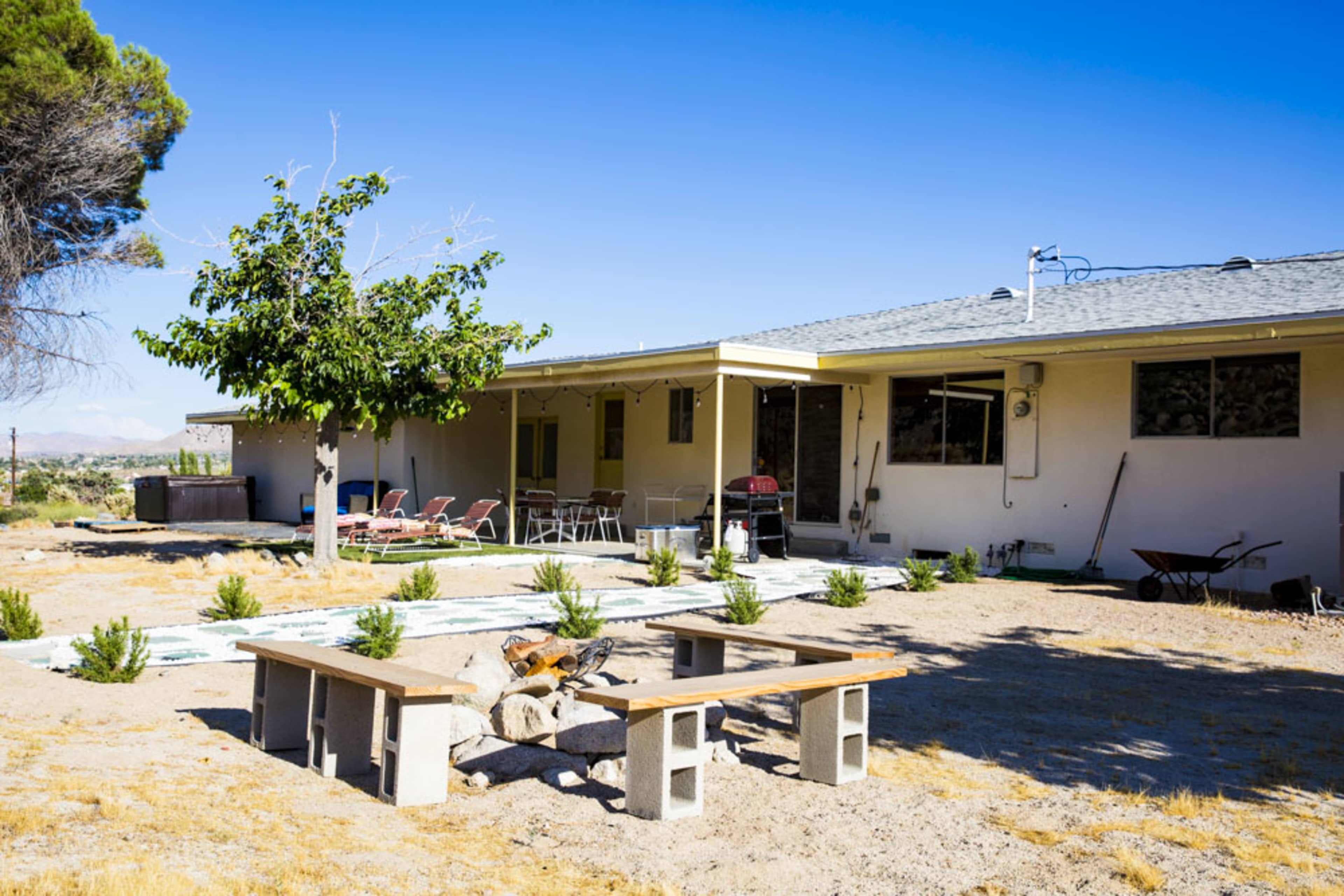 The image shows a single-story house with a landscaped yard featuring a stone seating area and a tree.
