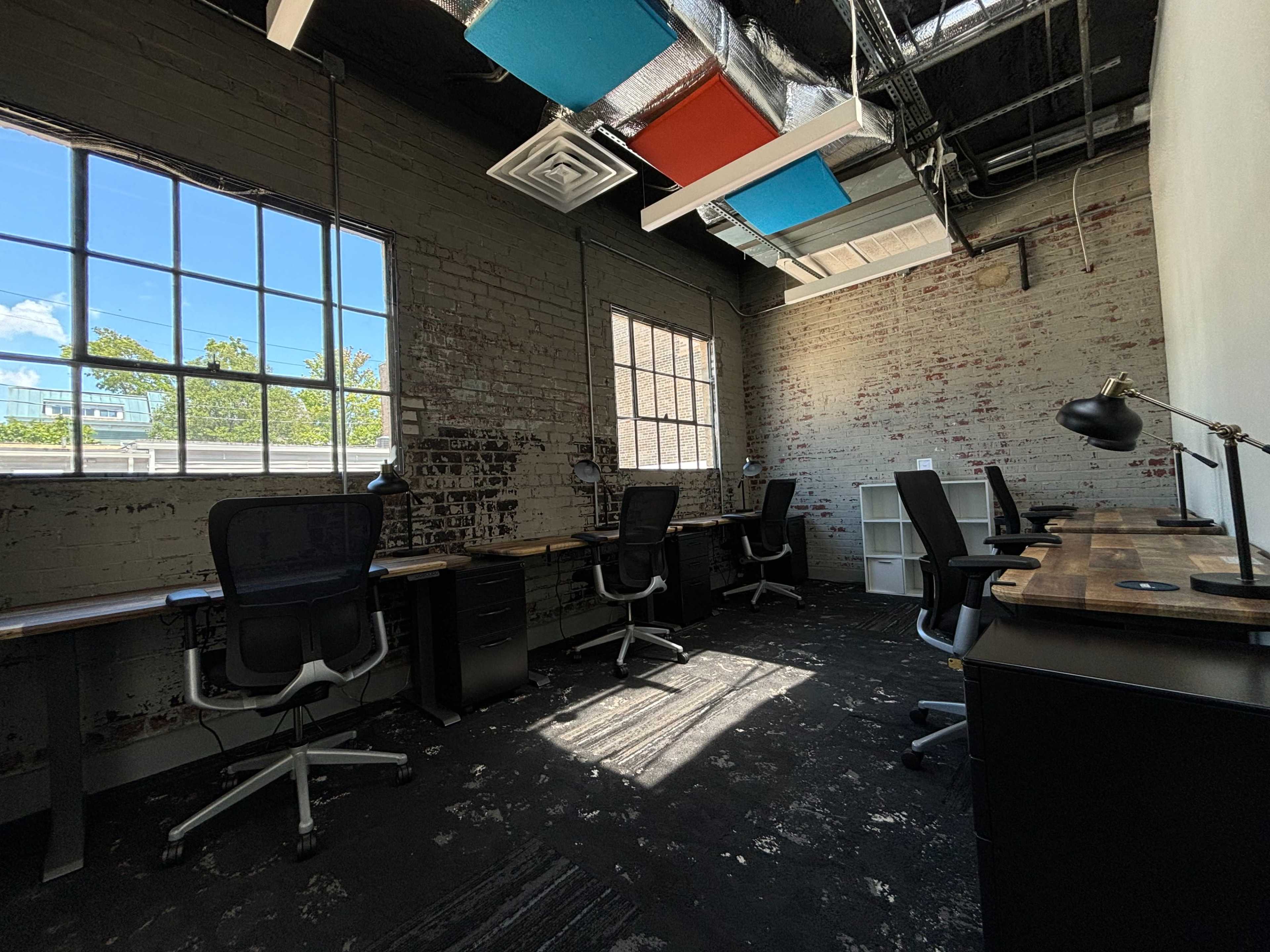 An office space with several desks, chairs, and large windows allowing natural light to enter.