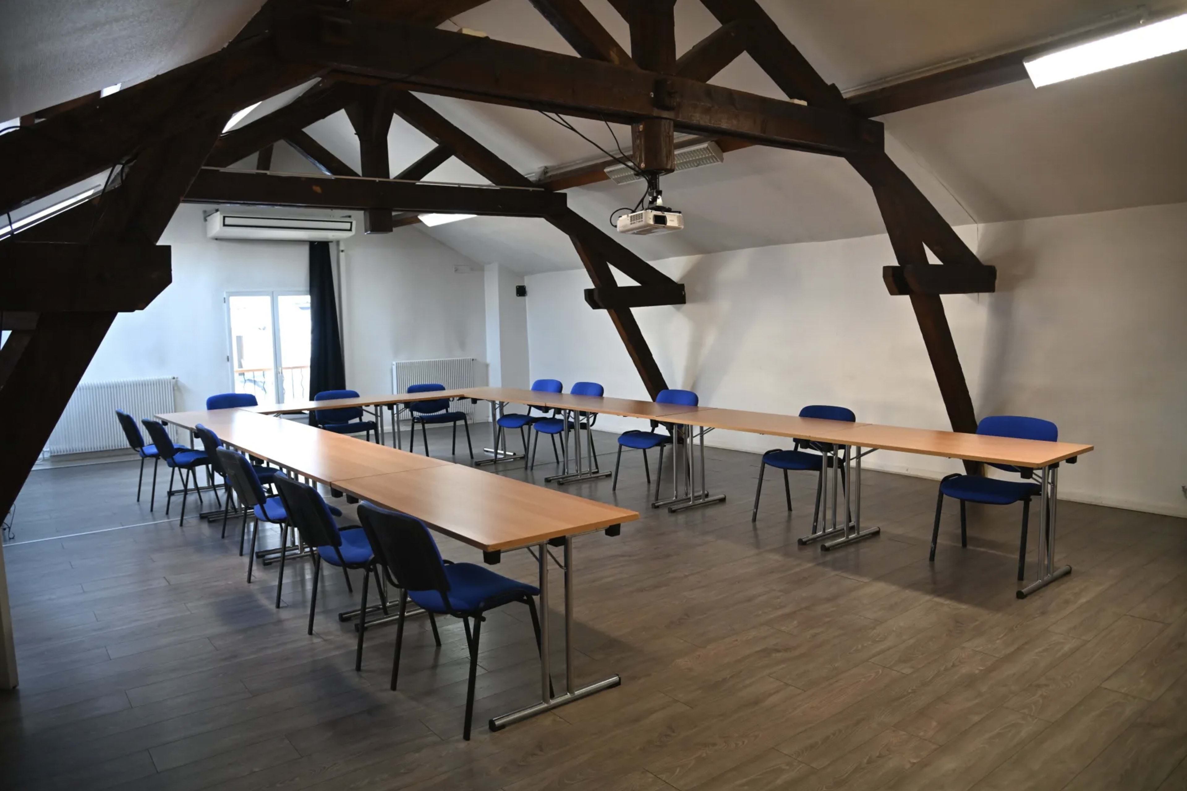 A spacious meeting room features several tables arranged in a U-shape with blue chairs, illuminated by natural light from a window.