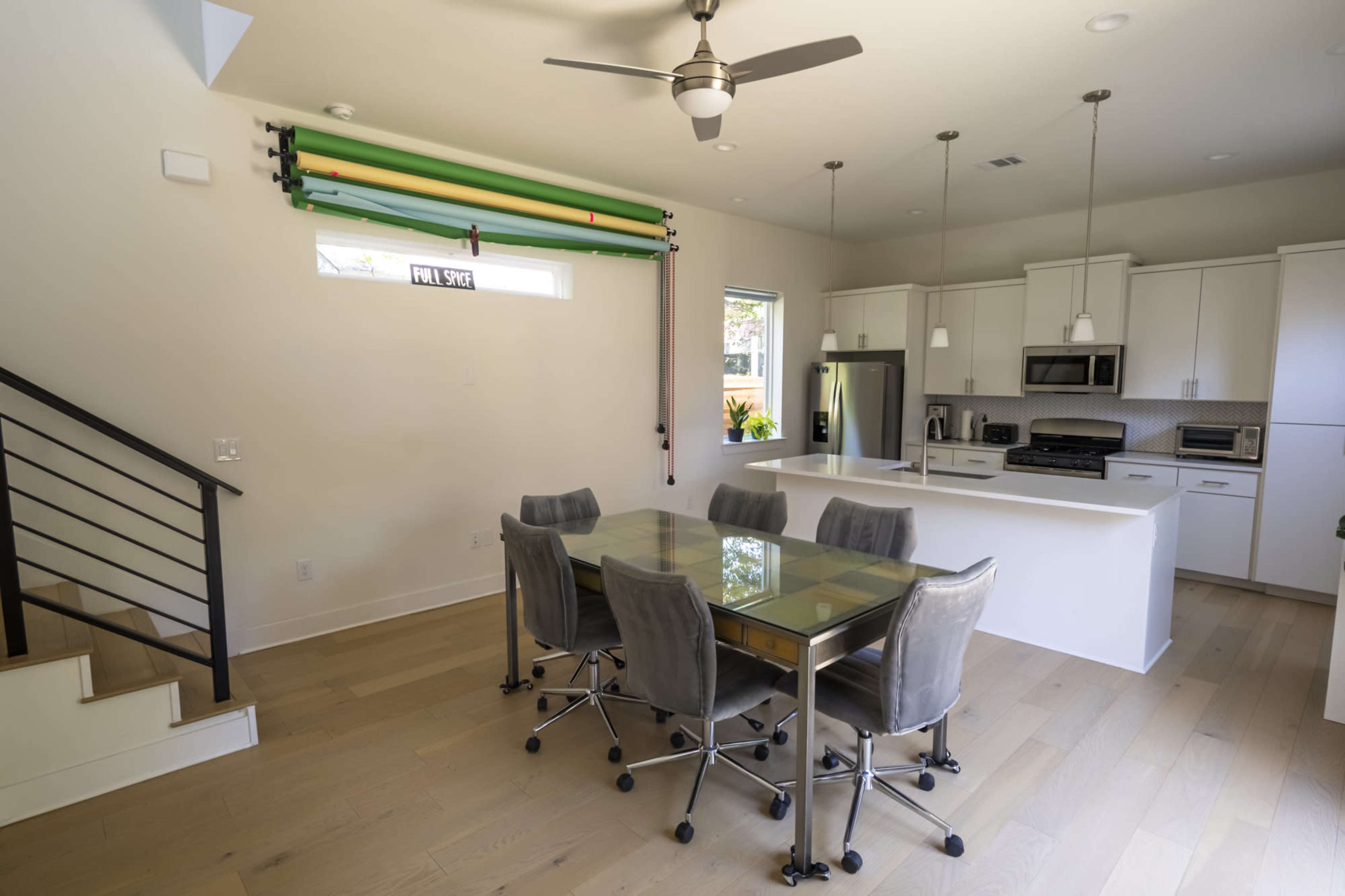 The image shows a modern kitchen and dining area with a glass table surrounded by wheeled chairs, stainless steel appliances, and a staircase on the left.