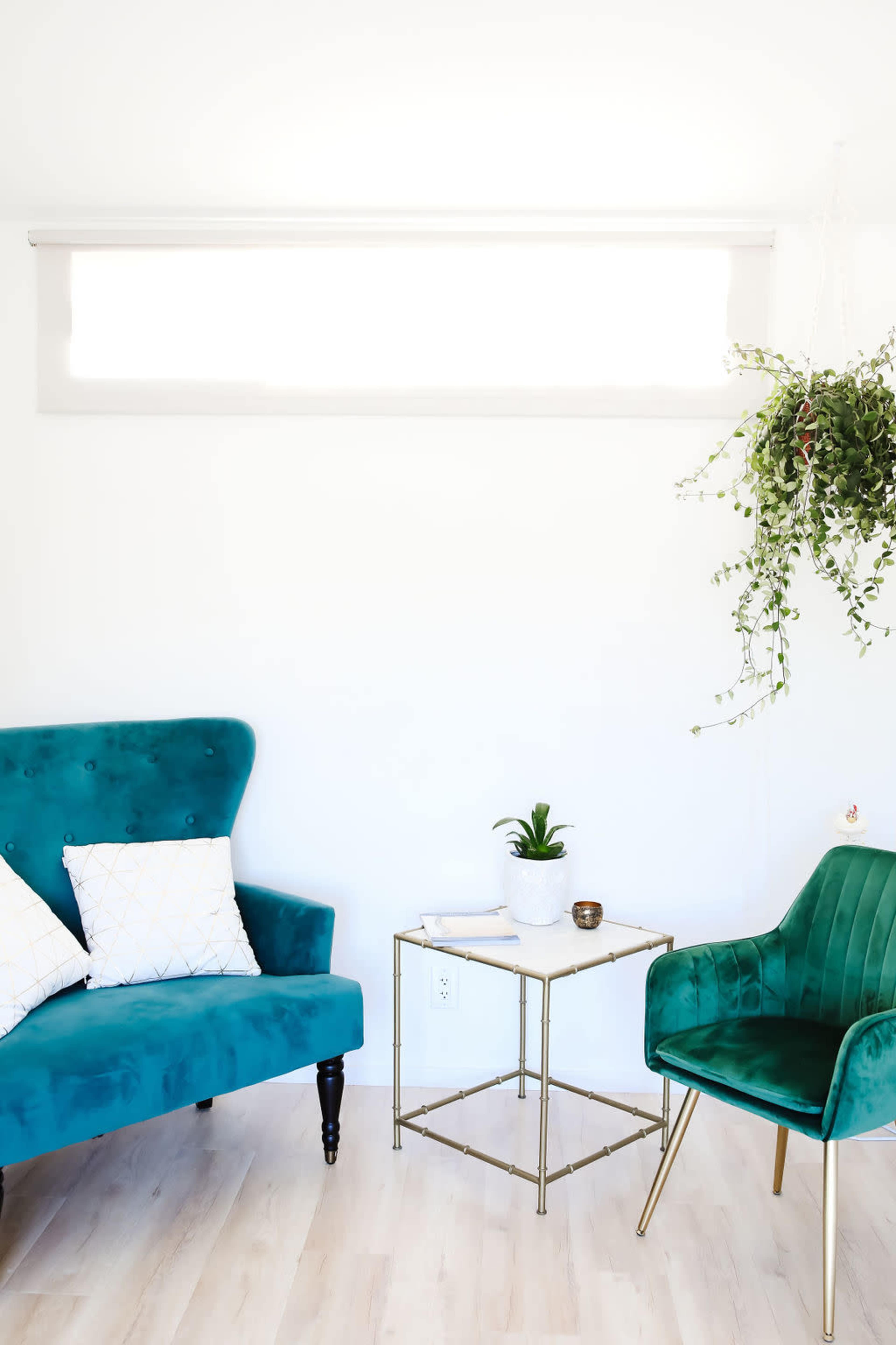 A bright, minimalist interior features a teal sofa and chair, a small table with a plant, and a hanging greenery accent.
