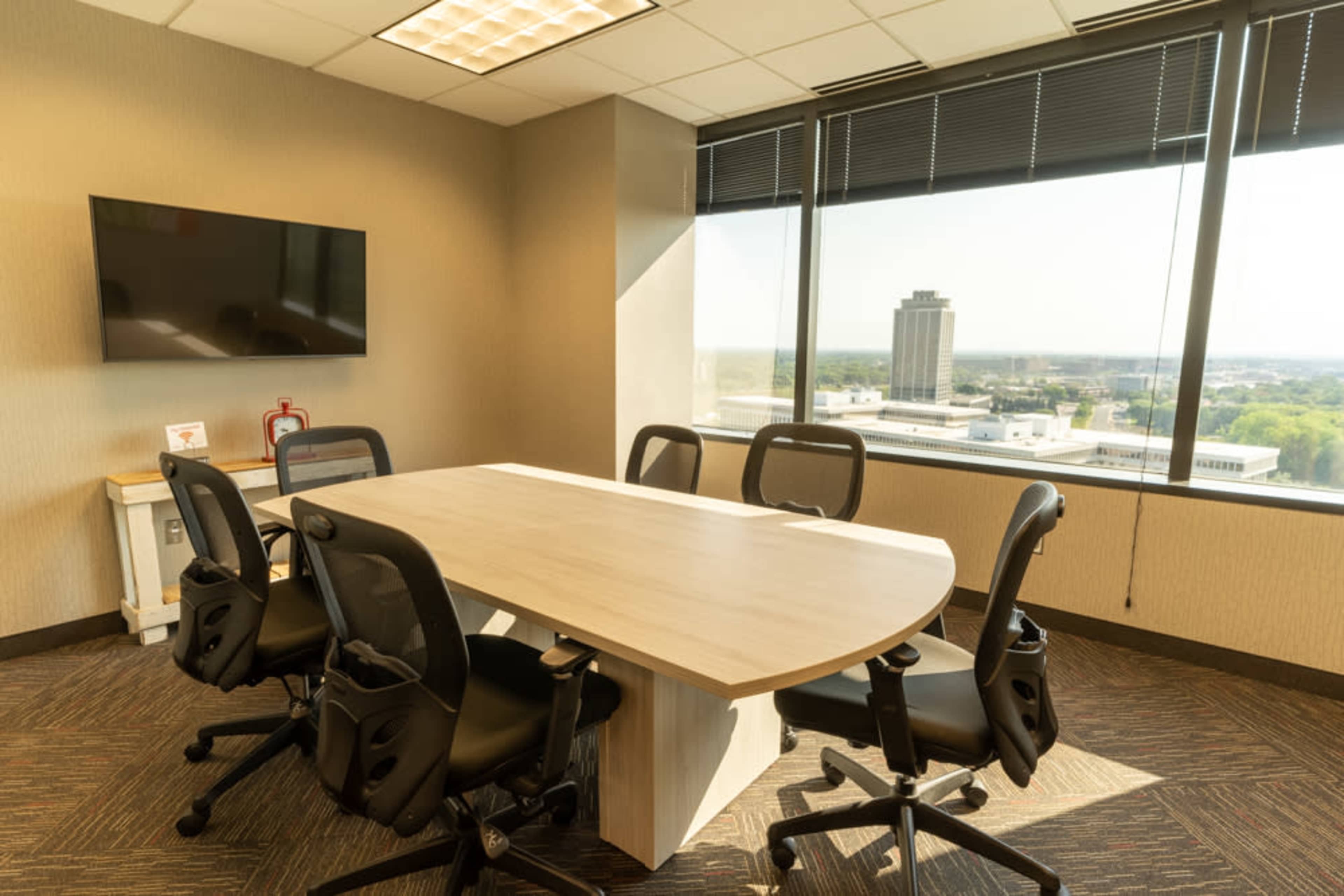 A conference room features a large oval table surrounded by six black chairs and a window with a view of a city skyline.