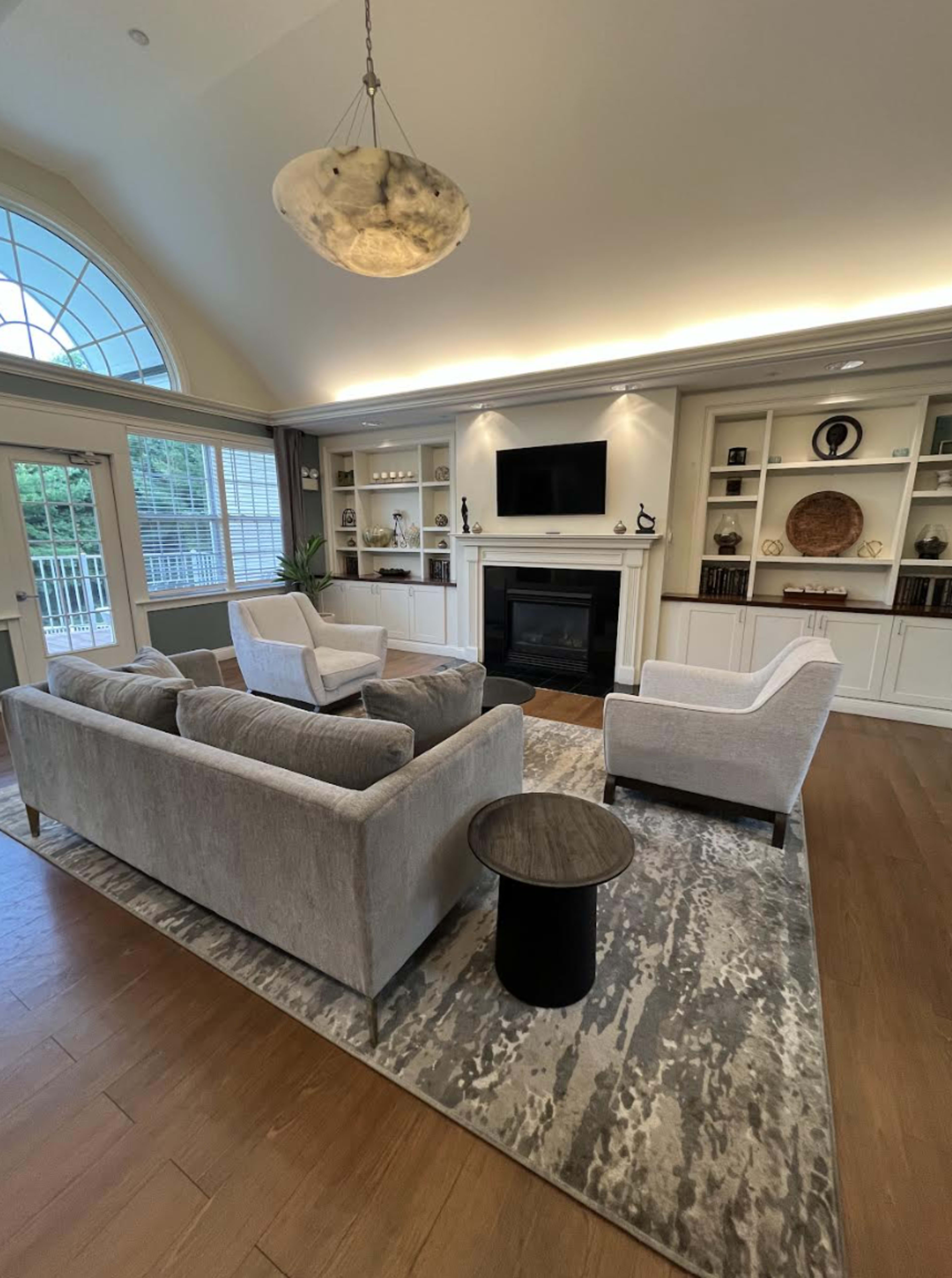 A modern living room features a gray sofa and armchairs arranged around a black fireplace, with built-in shelving and a large window allowing natural light.