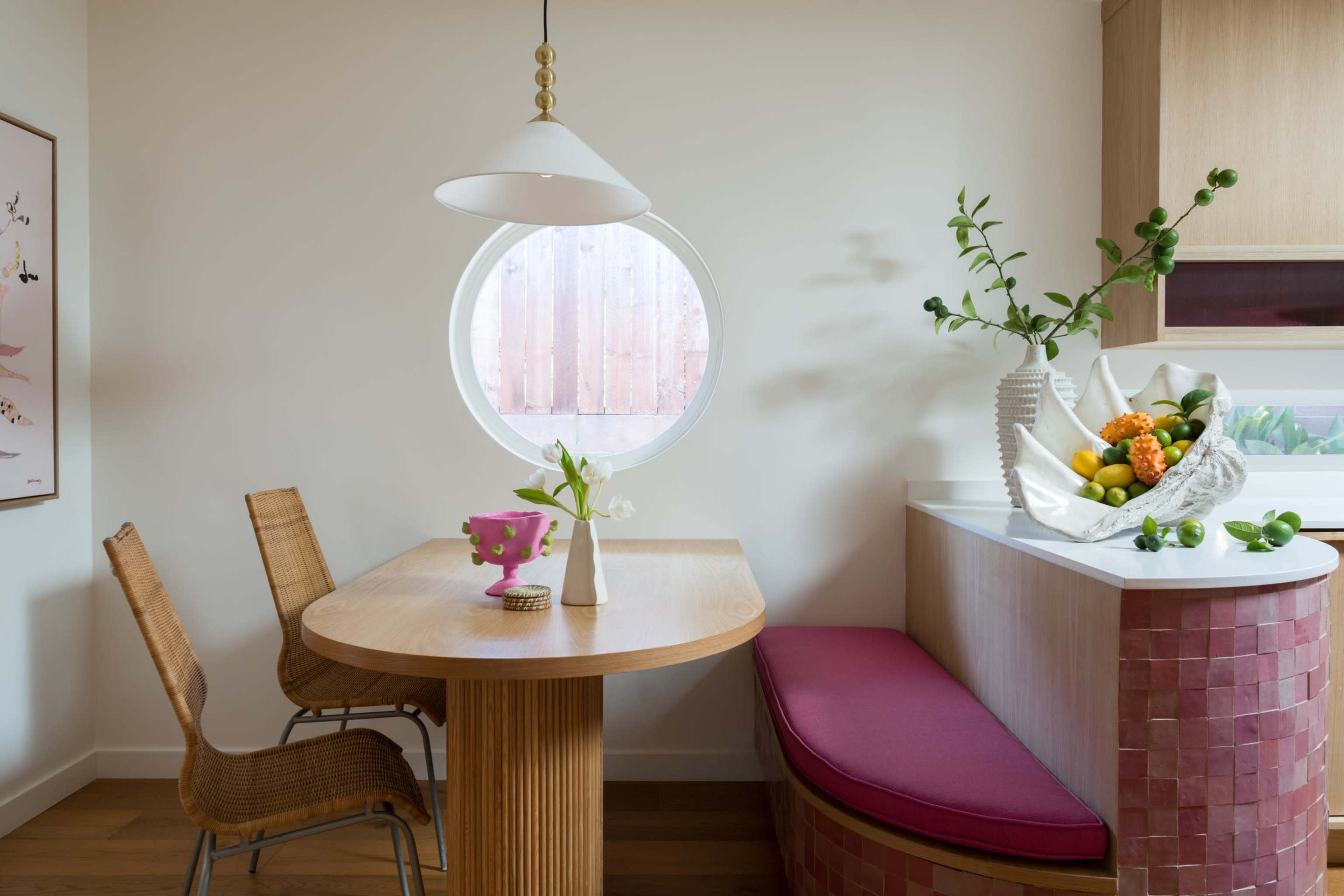 A small dining area features a round window, a wooden table with two chairs, and a curved bench with a colorful fruit arrangement on the counter.