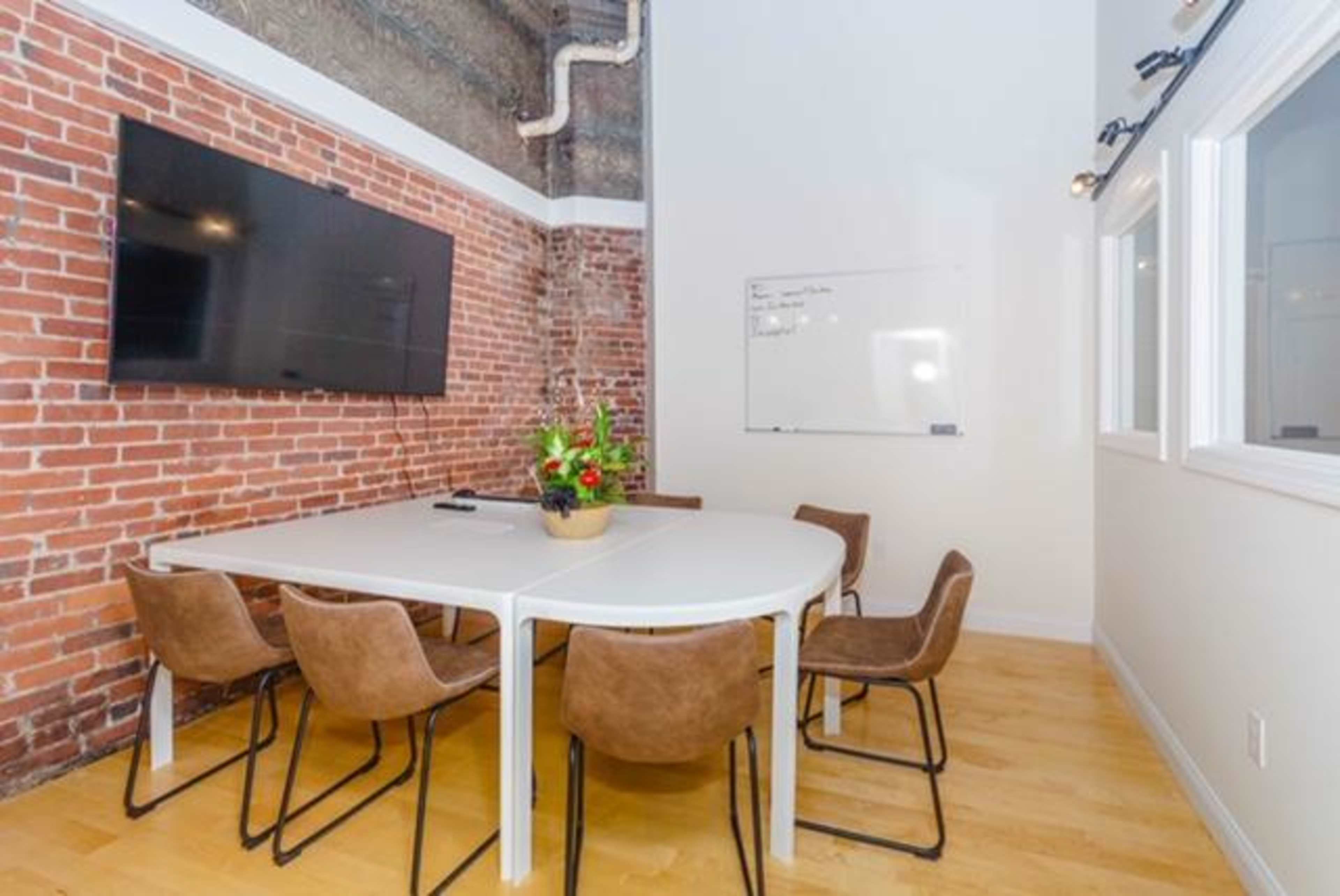A conference room features a round table surrounded by brown chairs, a large wall-mounted TV, and a whiteboard on the adjacent wall, all set against a backdrop of exposed brick.