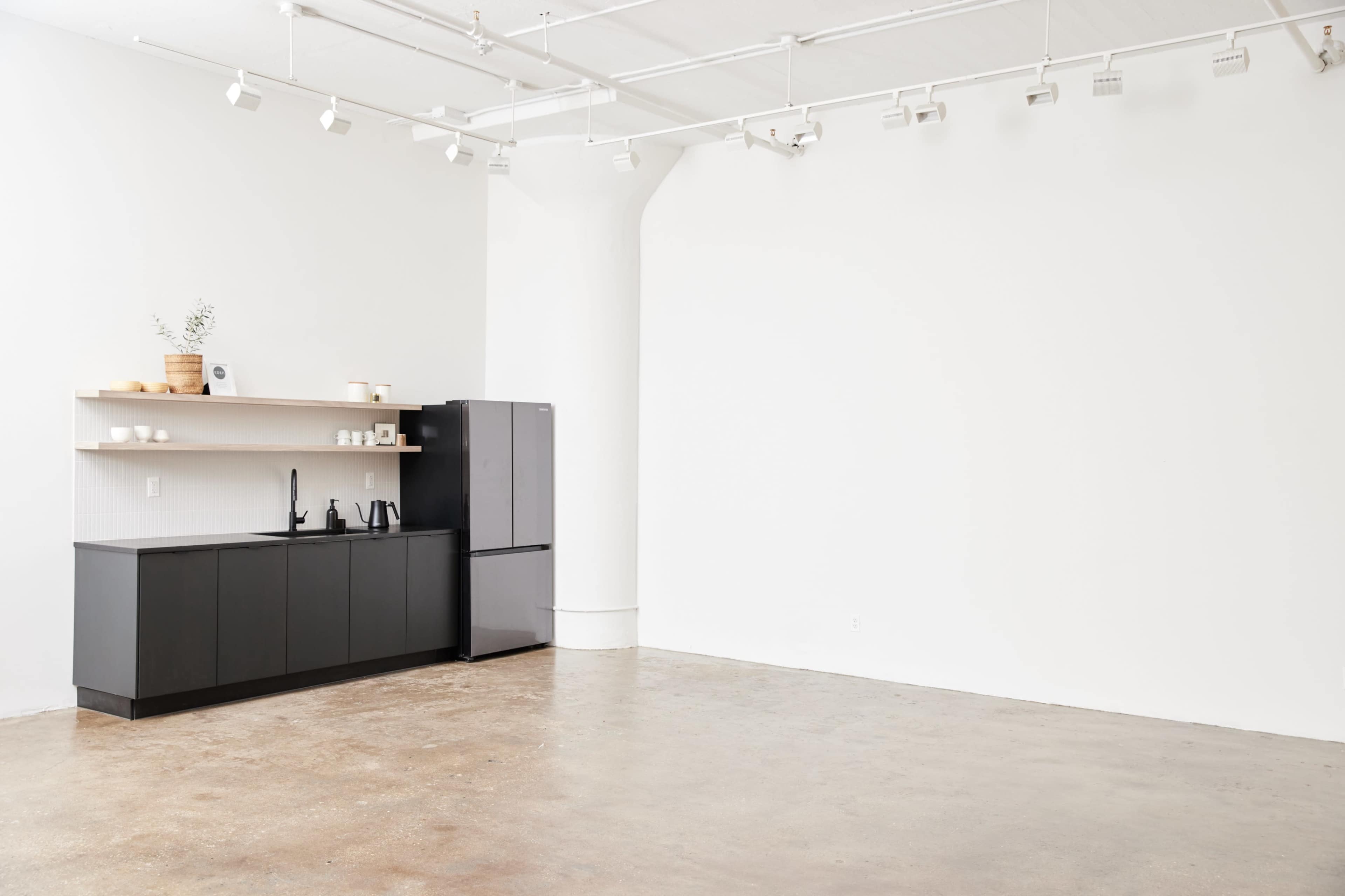A minimalist kitchen area with black cabinetry and a stainless steel refrigerator against a white wall in a spacious, empty room.