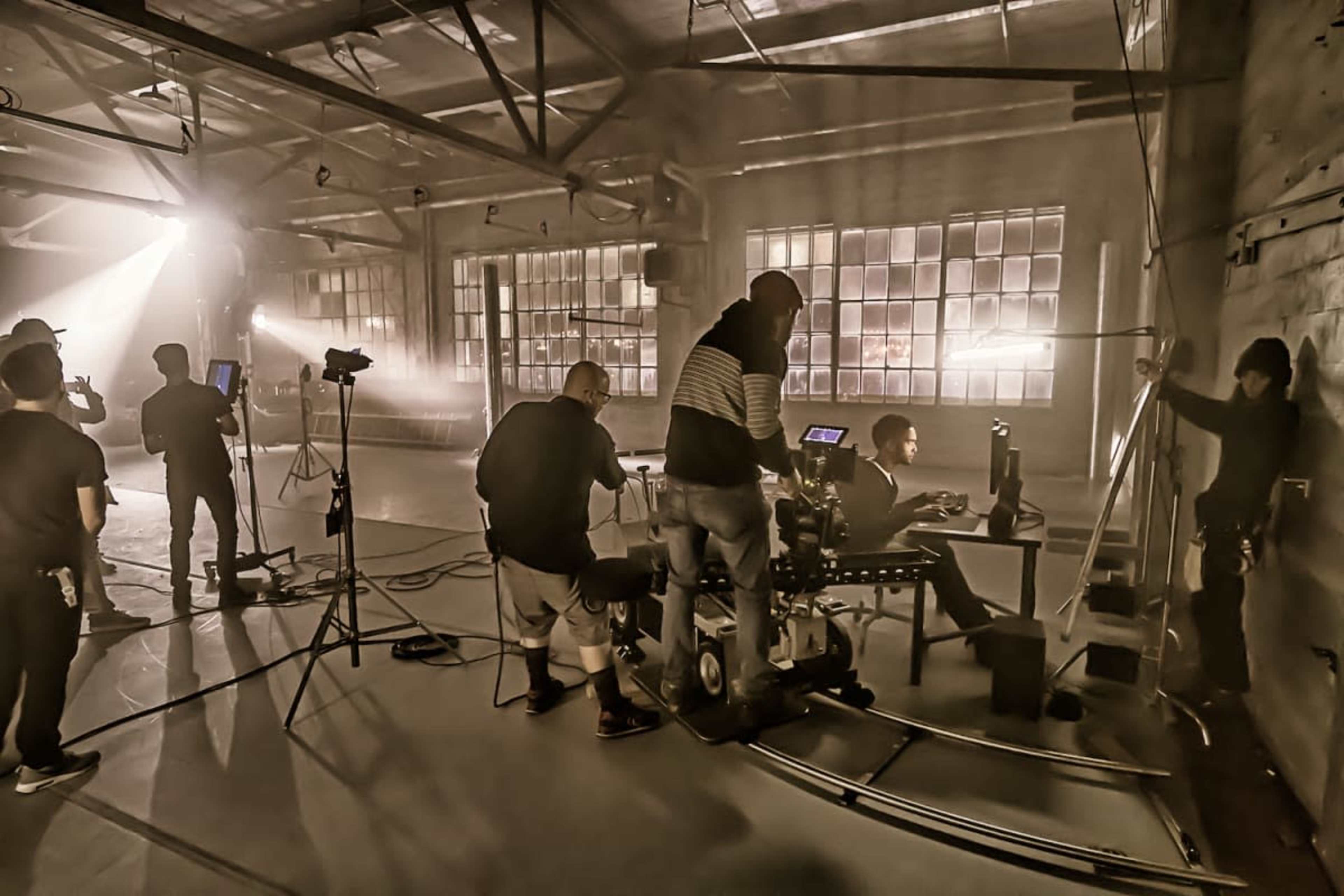 A film crew setups equipment and cameras in a dimly lit industrial space with large windows.