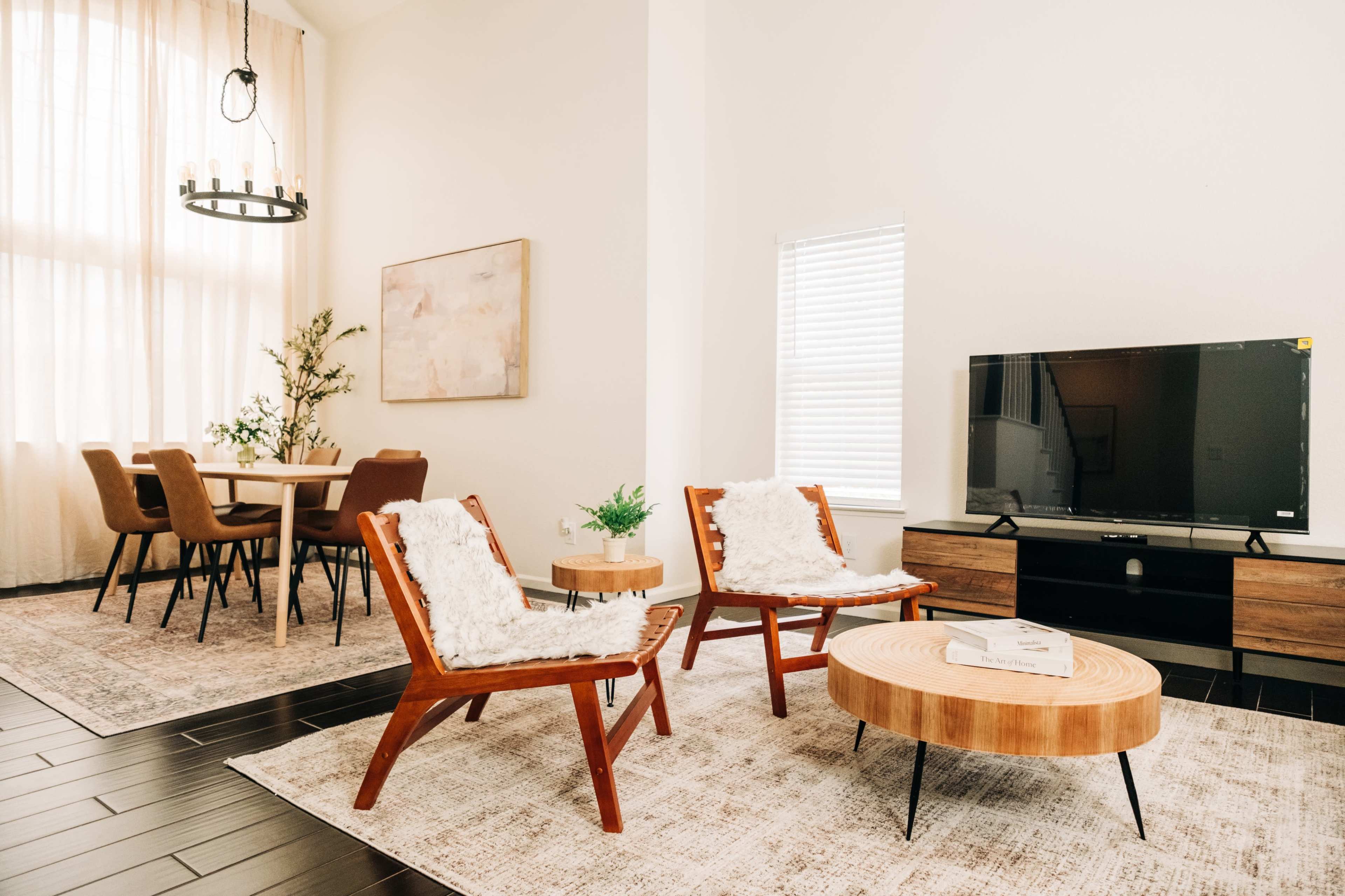 A modern living room features two wooden chairs with white cushions, a round coffee table, and a television on a low stand, alongside a dining area with a table and chairs.