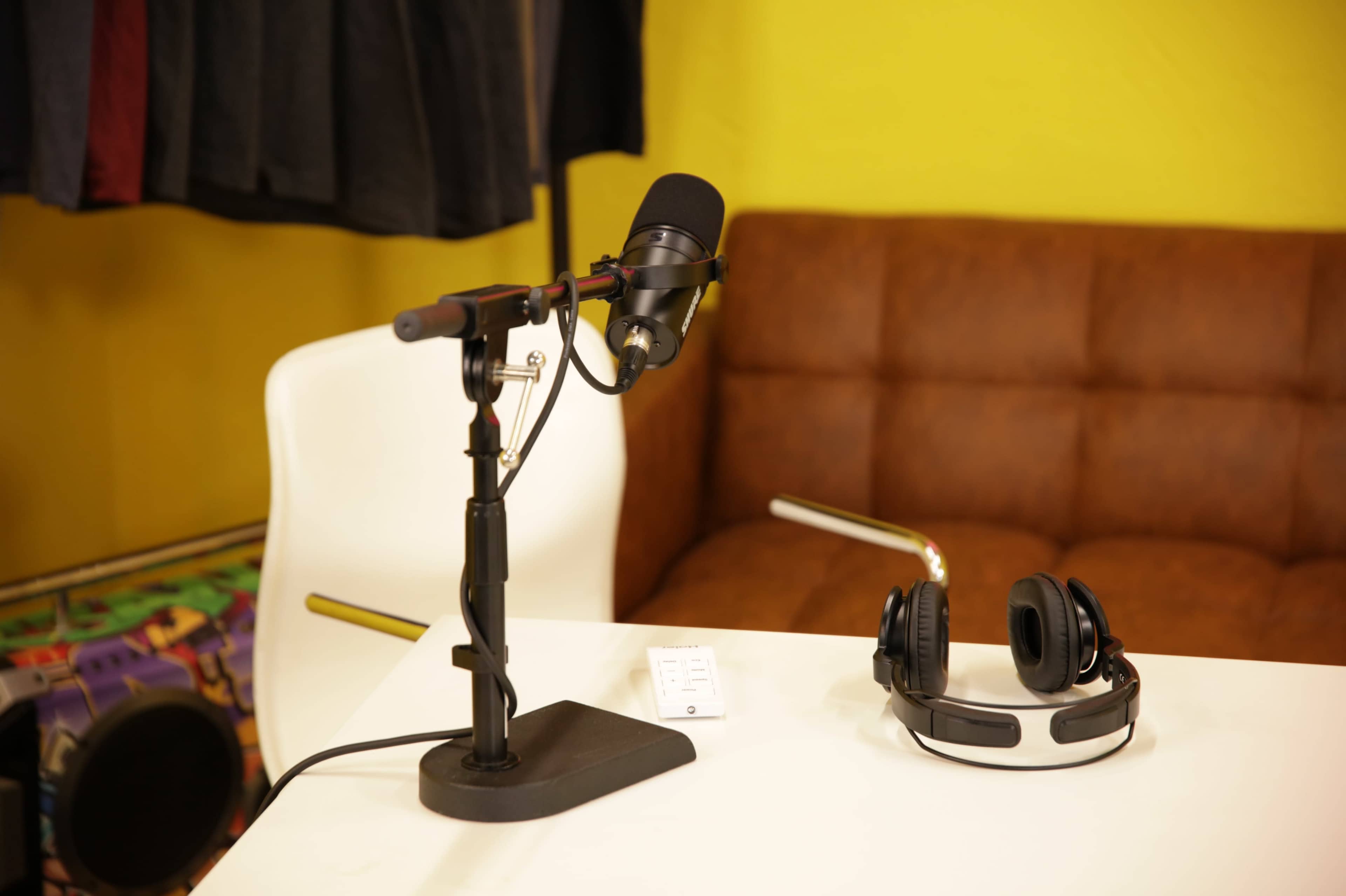 A microphone on a stand is positioned next to a pair of headphones on a table in a room with a yellow wall and a brown couch.